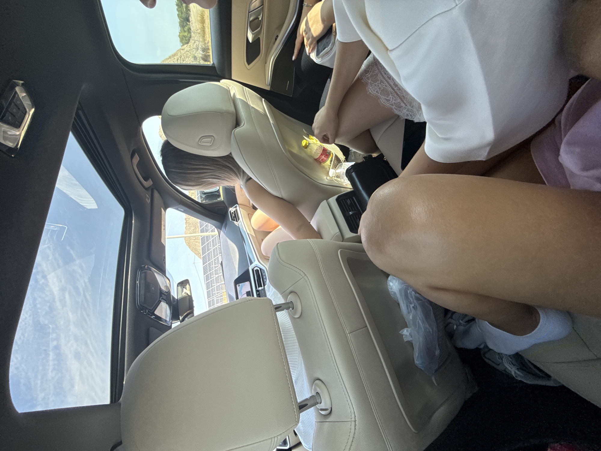 This is a photograph taken from inside a car, looking towards the rear passenger seats and out the windows. The car appears to be moving, as indicated by the dynamic view of the landscape outside. The interior of the car is visible, with beige leather seats and a sunroof overhead, revealing a bright, cloudy sky.
In the foreground, the legs and lower torso of at least two individuals are visible. One person is wearing a white garment, possibly a dress or robe, with lace trim. Another person has bare legs and is wearing white socks. Bags are visible on the floor of the car.
In the middle ground, the back of a head and shoulders of a woman with blonde hair is seen, sitting in a rear seat. She is looking towards the front of the car, and her arm rests on the seat. A bottle of water is visible in a seat pocket behind her.
Outside the car, a landscape of rolling hills or mountains is visible, along with what appears to be a solar panel farm in the distance. The environment suggests a rural or semi-rural setting. The presence of solar panels might indicate a developed area with renewable energy infrastructure. The bright sunlight and clear sky suggest it is daytime, likely warm weather given the attire of the people inside. The overall impression is of a road trip or journey through a scenic area. The location of Cârțișoara, Romania, is mentioned in the prompt, suggesting this scene is taking place in or around that area.
