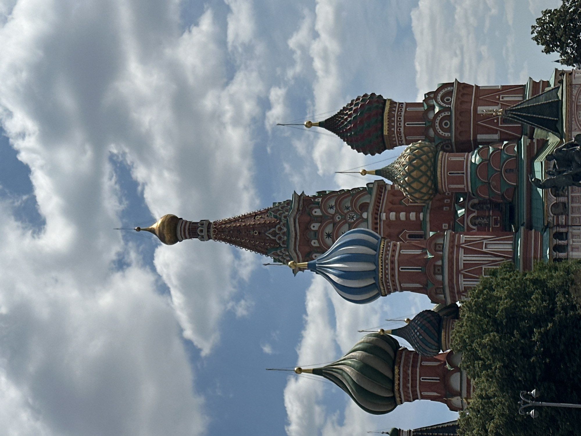 The image captures a close-up view of Saint Basil's Cathedral in Moscow, Russia, under a cloudy blue sky. The iconic onion domes of the cathedral are prominent, showcasing their vibrant colors and intricate patterns. Some domes are topped with golden finials. The red brick walls of the cathedral are visible, decorated with white accents and architectural details. Lush green trees frame the lower part of the structure, and a lamppost is visible on the left. The scene is static, with no people or overt actions depicted, focusing solely on the architectural grandeur of the cathedral against the backdrop of the sky.