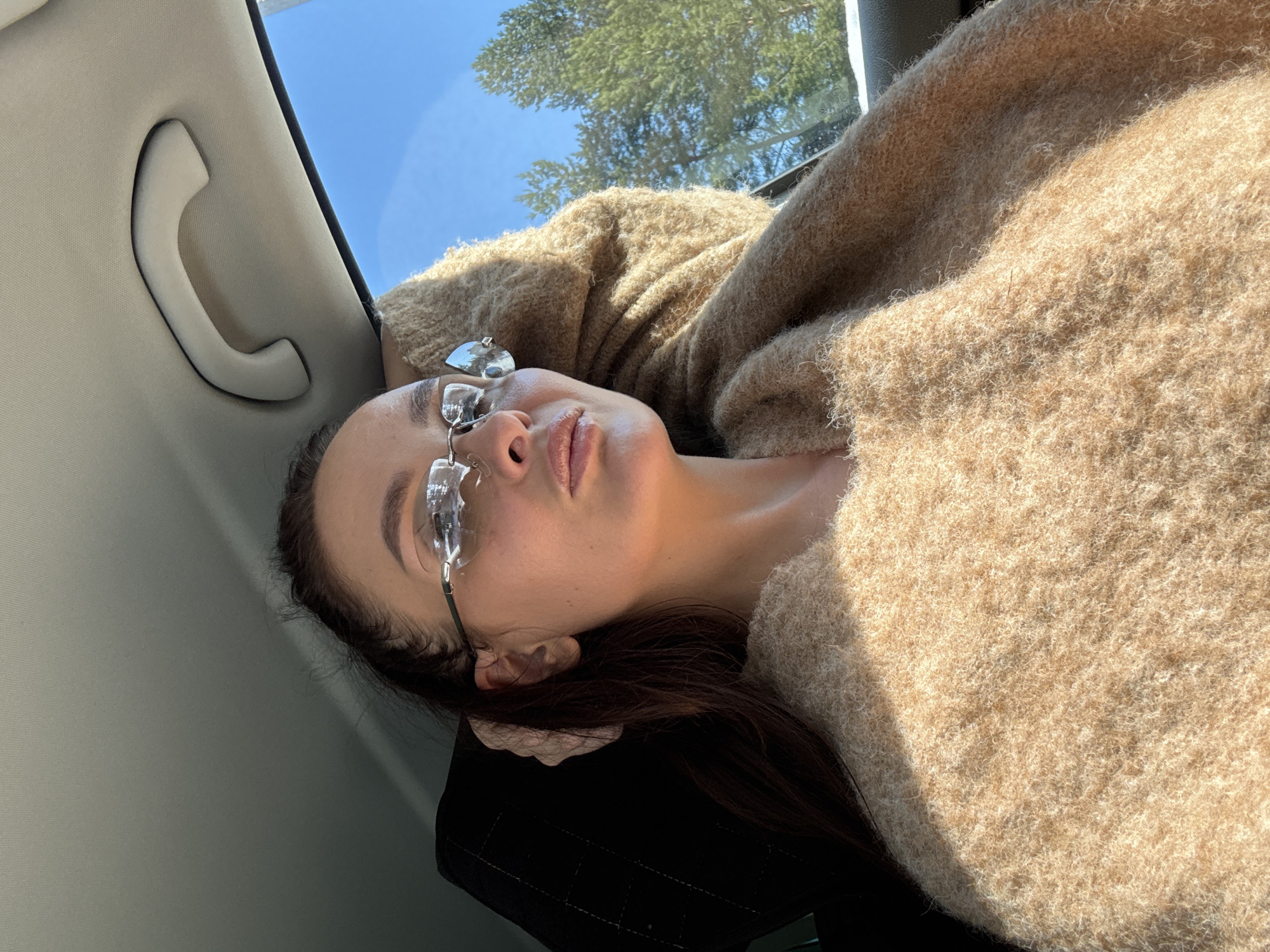 A woman lies in the back seat of a car, wearing a beige fuzzy sweater and glasses with dangling embellishments. She appears to be looking out the car window, where trees and a blue sky are visible. The car's interior is light gray, with a handle visible above her head. There is no evidence that this image was taken in Chaikovsky, Russia.