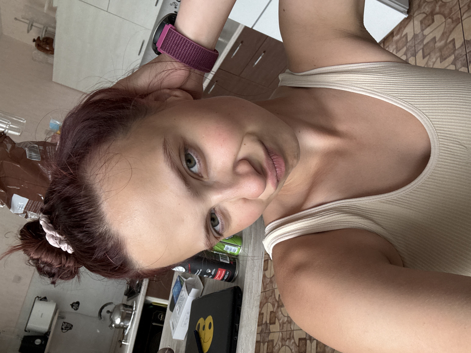 A person is in the Прикамский, Russia area, indoors. The individual has reddish-brown hair pulled back into a bun, green eyes, and is wearing a light-colored ribbed tank top. A fitness tracker is visible on their wrist. They appear to be in a kitchen-like area with cabinets, appliances, and assorted objects like food containers, cans, and a laptop or tablet with a smiley face sticker on it. The floor is tiled.
