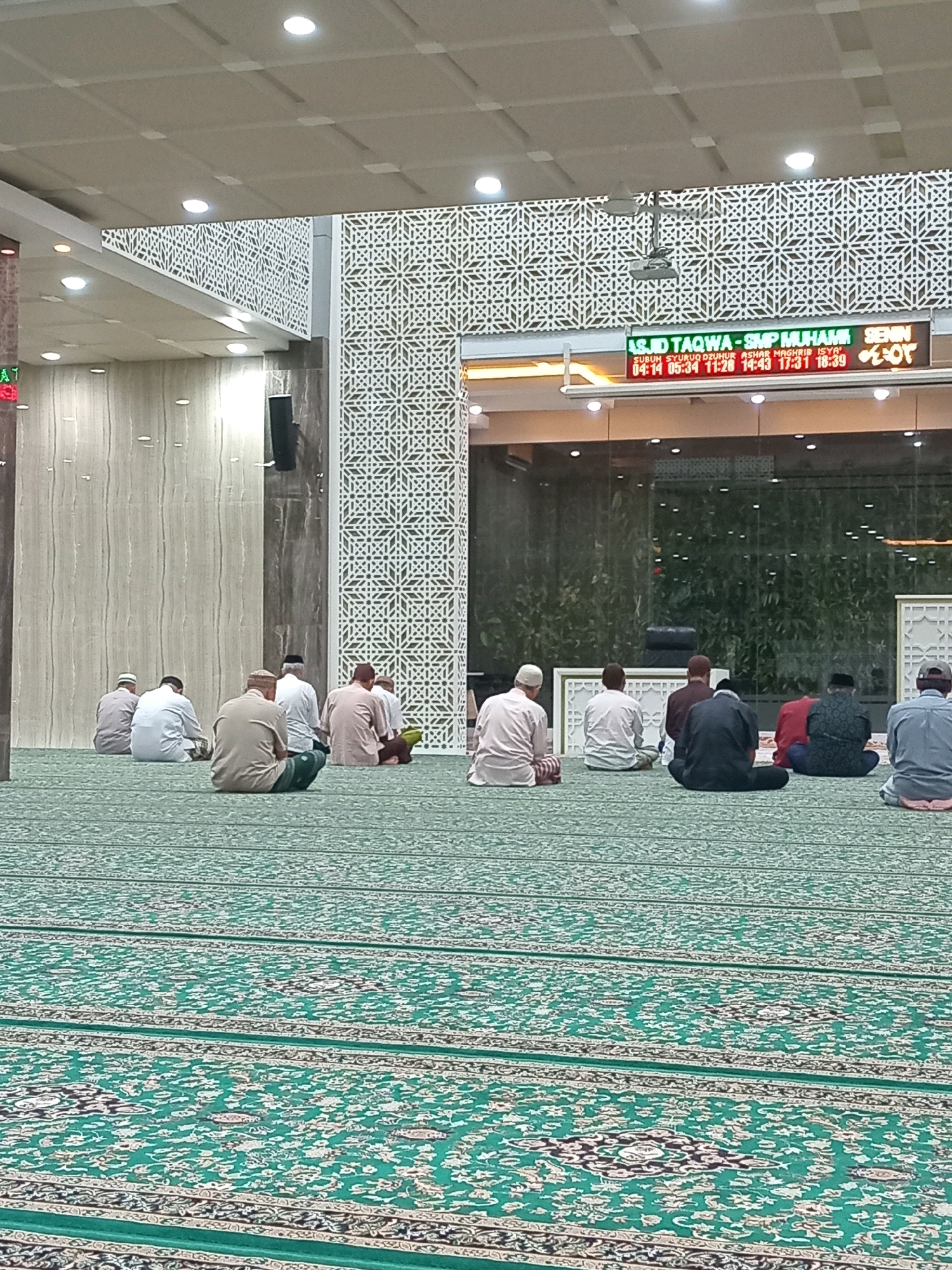 The image depicts the interior of a mosque, specifically the Masjid Taqwa - SMP Muhammadiyah in Randuagung, Indonesia. Several men are seated on the green carpeted floor, facing a glass wall or window that looks out onto an indoor greenery. The men appear to be engaged in prayer or religious contemplation. The mosque features modern architectural elements, including a patterned screen on the wall and a digital display showing prayer times. The lighting is bright and even, illuminating the entire space.