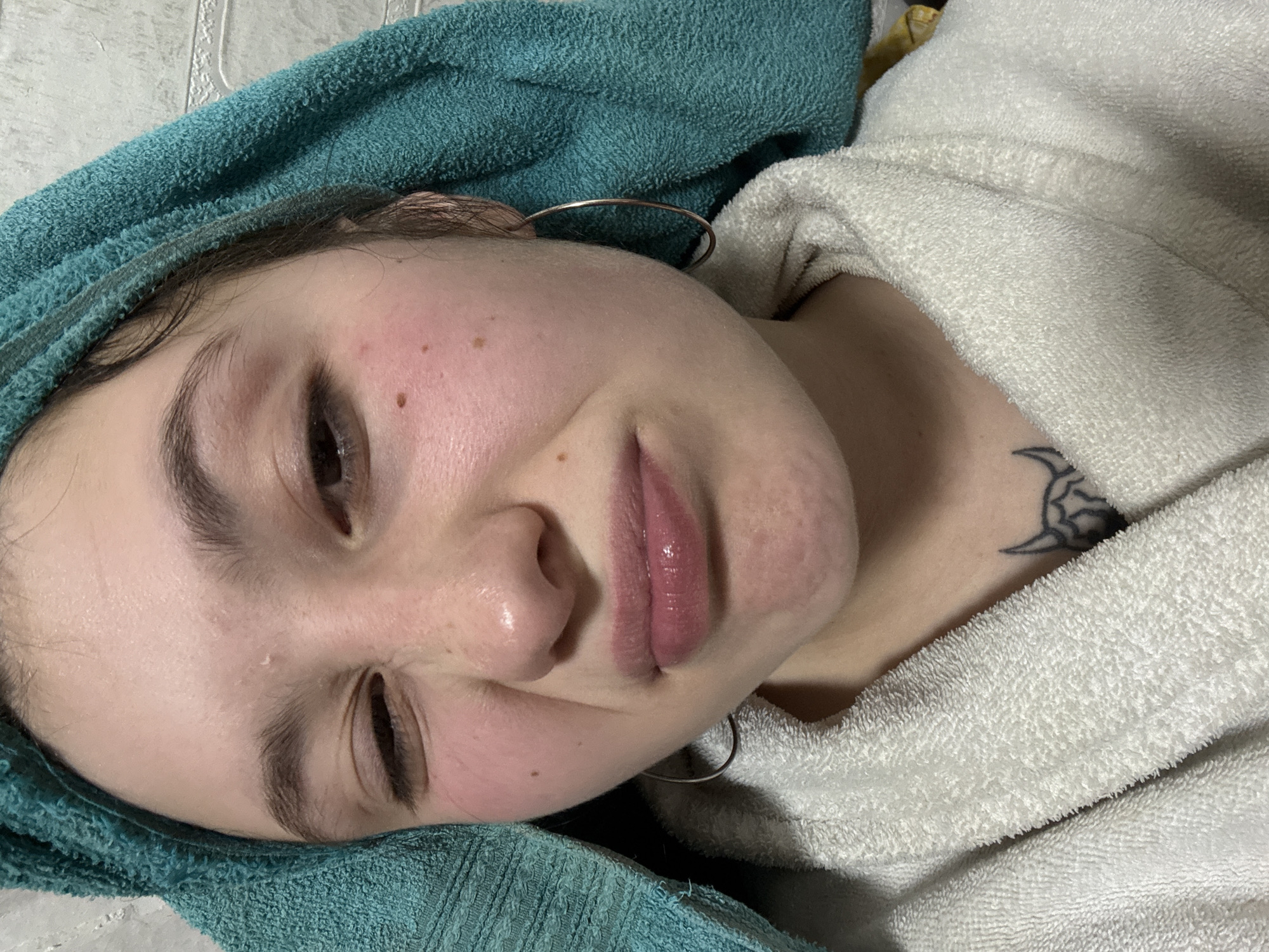 A close-up shot depicts a young woman with fair skin, wearing a light-colored, plush robe. Her head rests on a green towel. A pair of thin, silver hoop earrings are visible. On her chest, above the robe's collar, is a black tattoo. Her eyes are partially closed. The scene appears to be indoors. The exact location within the city of Фоки, Russia is not identifiable from the provided image.