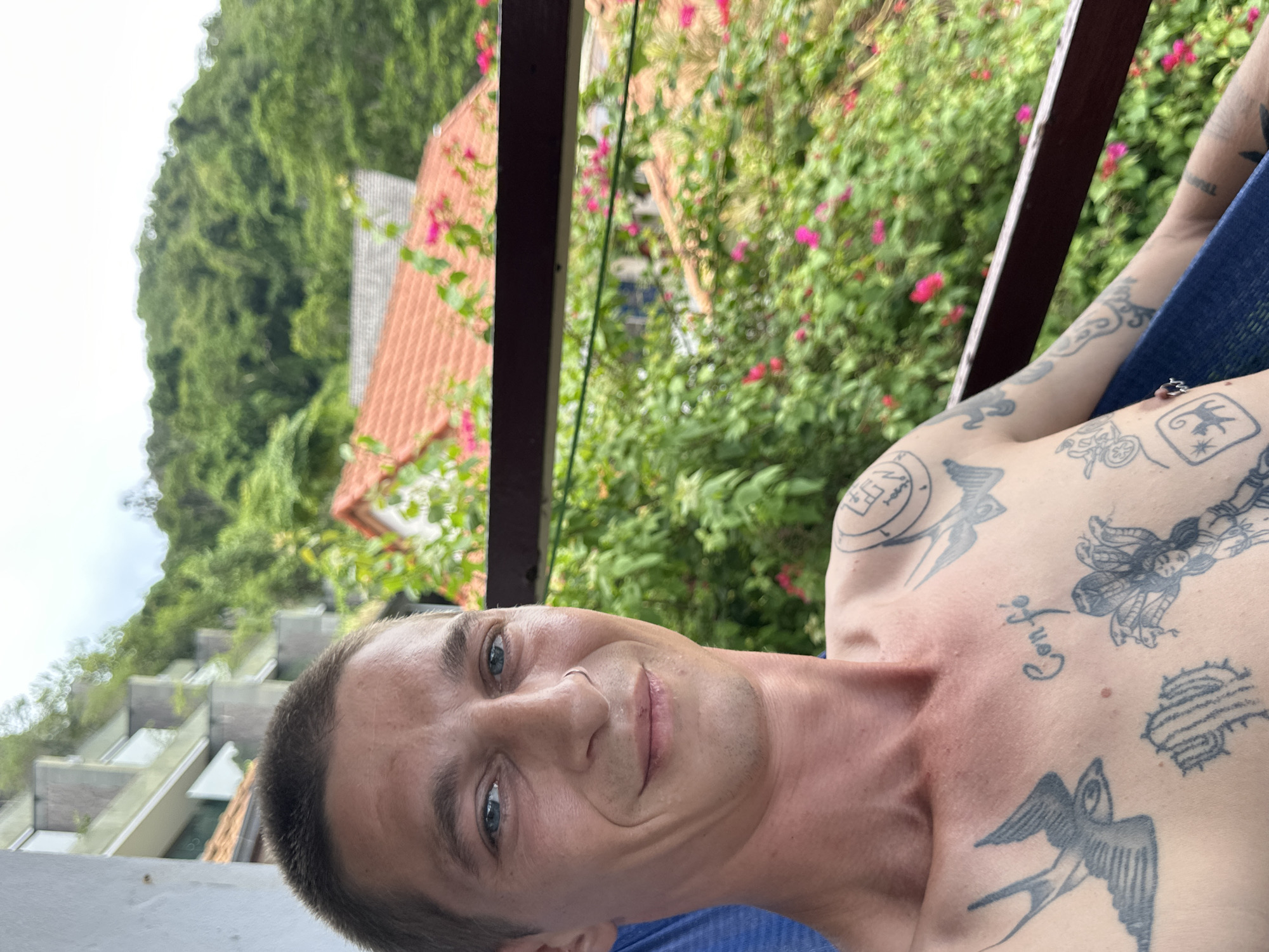 A man with short, dark hair and blue eyes is pictured from the chest up, sitting outside. He is wearing a silver hoop in his nose and has numerous tattoos on his chest and arms, including swallows, a cactus, and a female figure with snakes for hair. He is smiling slightly and looking towards the camera. The background shows lush green trees and foliage, with some pink flowers visible. Behind the foliage, there are buildings with orange tiled roofs, and a multi-story building with concrete balconies. The scene appears to be a balcony or outdoor seating area overlooking a tropical landscape. The location is identified as Karon, Thailand.