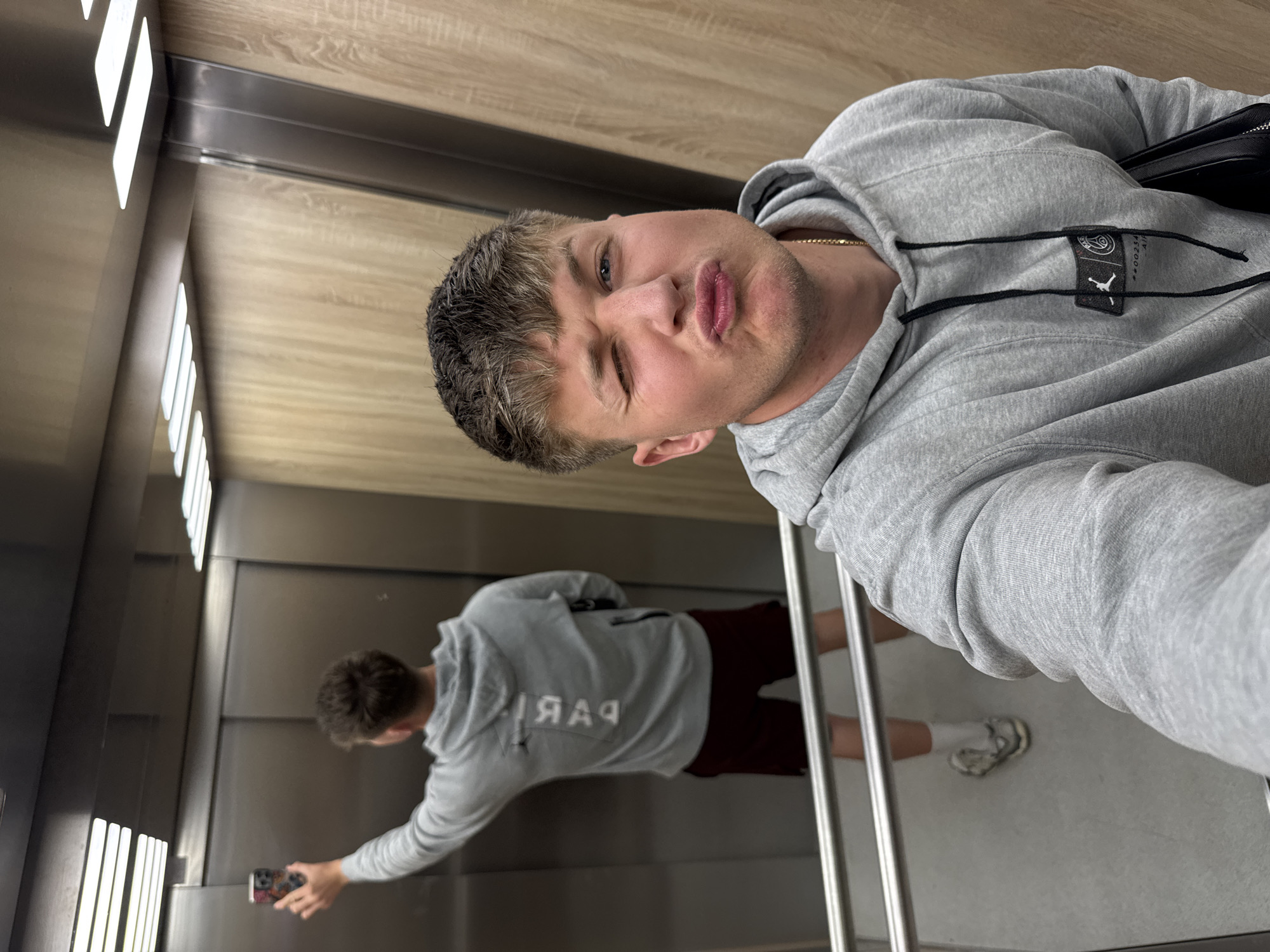 A man in a grey hoodie with the word "PARIS" reversed on the back is taking a selfie in an elevator with the reflection of himself taking the picture. The elevator has wooden paneling at the top and mirrored panels on the walls. The man is wearing a gold chain and has a black bag slung over his shoulder. He is pouting and winking at the camera. The location is an elevator in Plovdiv, Bulgaria.