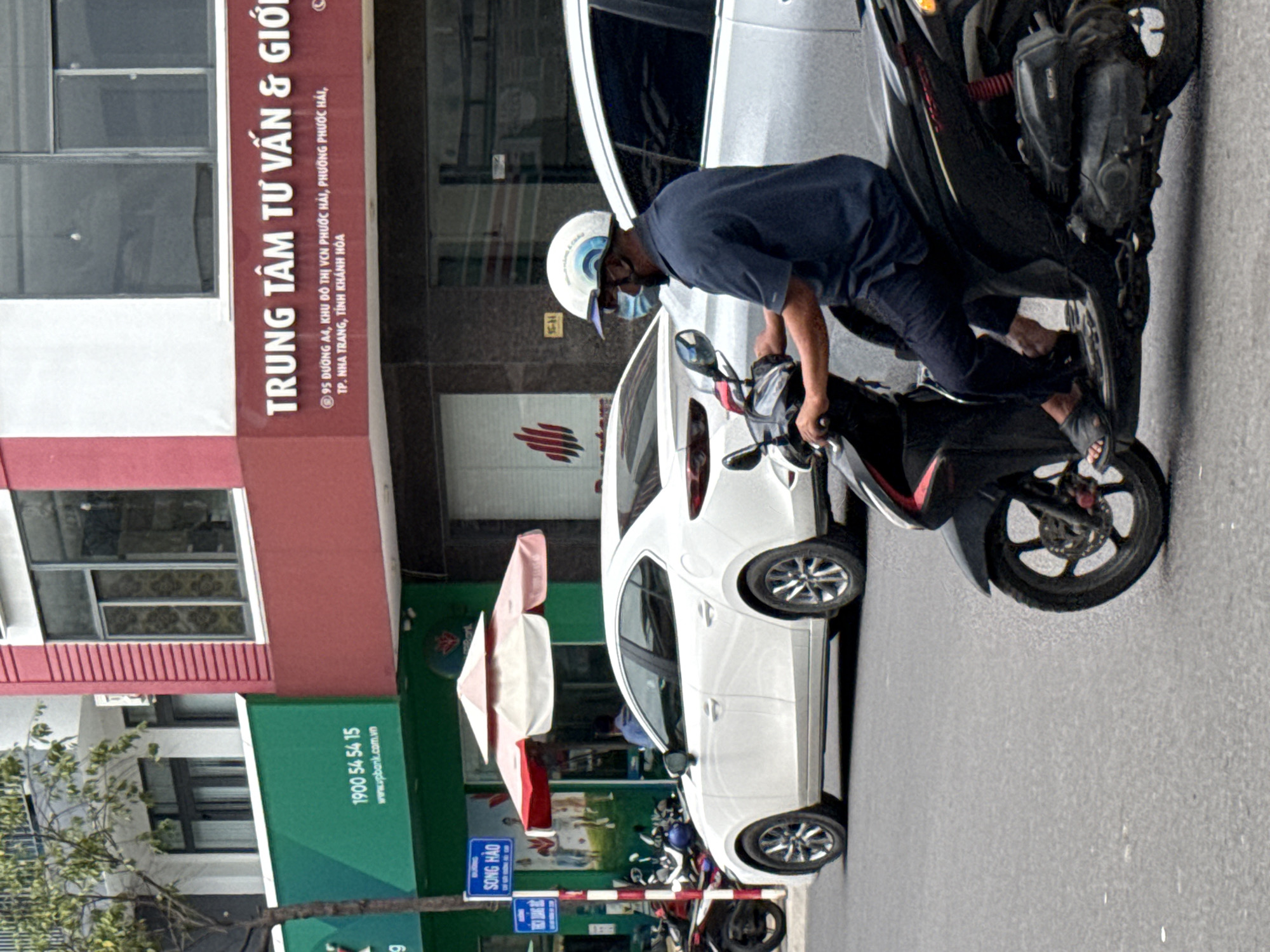 The image depicts a street scene in Nha Trang, Vietnam. In the foreground, a man wearing a dark blue shirt, dark pants, and a white helmet with a blue wave pattern is riding a black scooter. He is also wearing a light blue face mask. The scooter has red accents on the front panel. Behind the scooter, a white sedan is parked, and further back, another white car is visible. The background features a building with a red and white facade. A green sign with Vietnamese text and a phone number "1900 54 54 15" is mounted on the building. Another sign with Vietnamese text is above it. A tree with green foliage is partially visible on the left side of the image. The street is paved with asphalt.