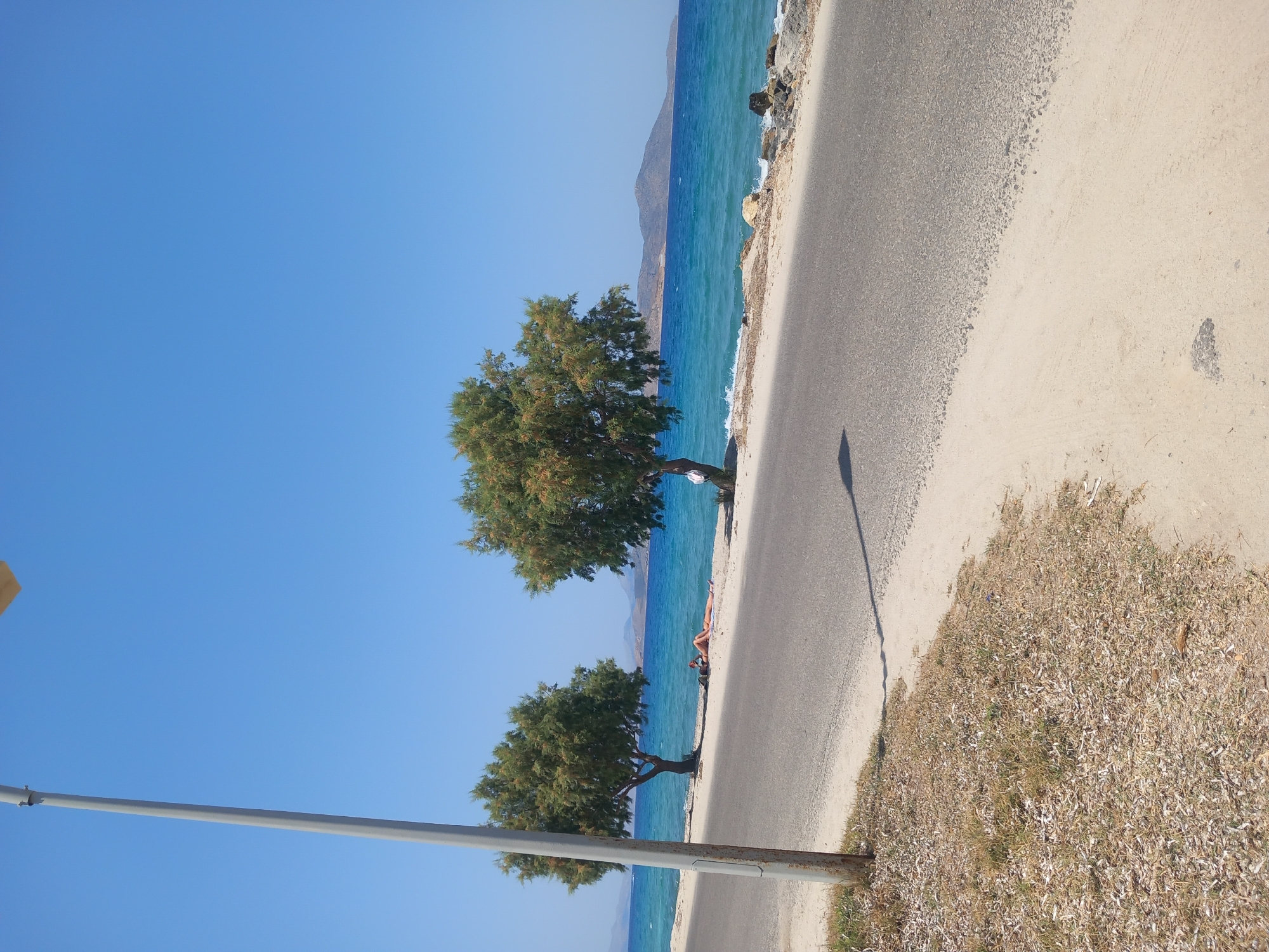 A street runs parallel to a body of water, with two trees and a person sunbathing on the sandy shore between the street and the water. The sky is clear and blue. The scene is likely in Zipari, Greece, a coastal town. The street is paved and shows tire marks. The sand is light in color and dry, with some patches of dry grass. The trees are green and leafy. The water is a bright blue, with small waves lapping at the shore. In the distance, hills can be seen on the horizon.