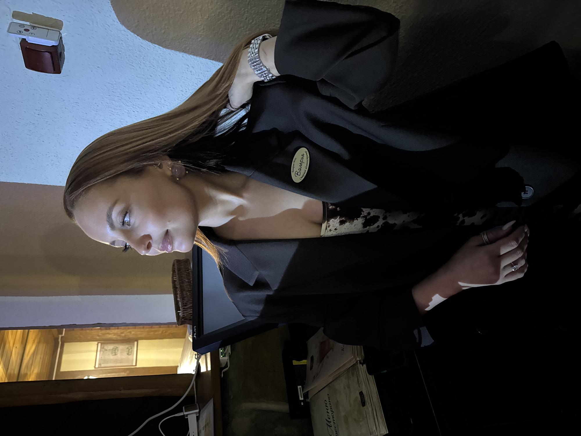 The image shows a woman with light brown hair wearing a black blazer and a bracelet, posing indoors. The location appears to be a restaurant or similar establishment in the city of Chaikovsky, Russia. This is indicated by the presence of a name tag on her blazer with the name "Валерия" and the Russian phrase "Оператор Абонентского отдела." There are menus in Russian visible in the background. The woman has her hand near a table or counter. In the background, there's a monitor, part of a wall with wooden paneling, and some equipment. There is also a ceiling-mounted object that could be a light fixture or sensor.