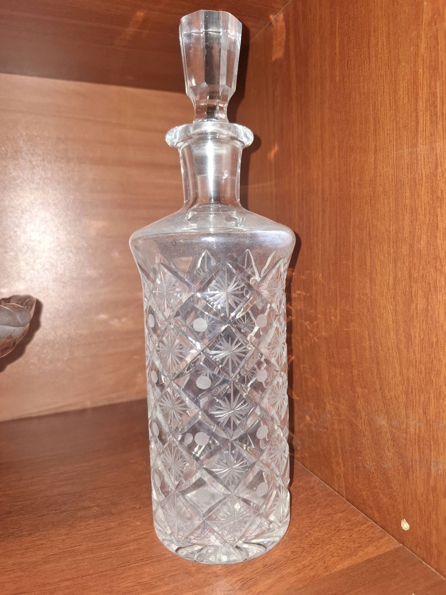 A crystal decanter with a geometric cut pattern and a stopper is displayed on a wooden shelf. The decanter is made of clear glass and has a faceted stopper. The shelf is made of polished wood with a visible grain pattern. The background is a wooden surface, likely part of a cabinet or shelf unit, with a warm, reddish-brown tone. There are no people or animals present in the image, and no discernible activity is taking place. The lighting suggests an indoor setting, possibly a home or display case. The image does not provide any information to determine its location in the city of Almaty, Kazakhstan.