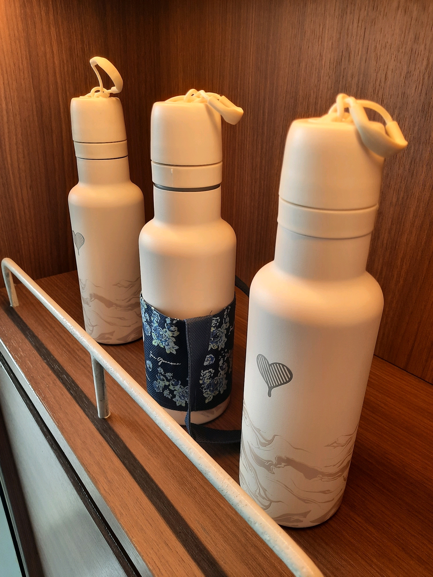 Three light-colored water bottles stand on a wooden shelf. The bottles are cylindrical with rounded tops and appear to be made of metal. The bottle on the far left has a faint heart design and a lighter gray marble-like pattern on its lower half. The middle bottle has a blue floral sleeve around its middle section. The bottle on the right also features a heart design and a marble-like pattern. A white metal railing is visible in the foreground, running horizontally across the image. The background is a dark wood-paneled wall. The scene suggests an indoor setting, possibly a hotel room or a retail display. The location in East Jakarta, Indonesia is not discernible from the image.