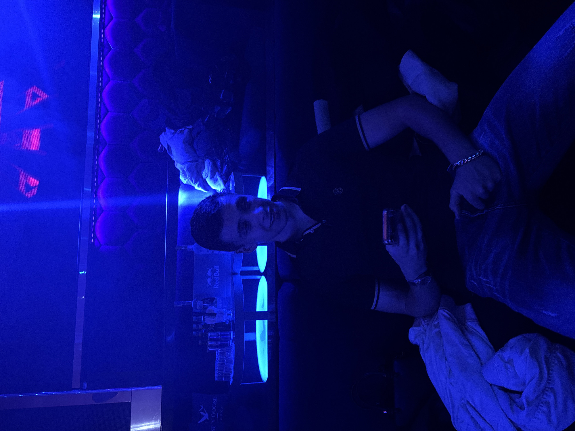 The image captures a young man in a nightclub or bar setting, characterized by predominant dark blue and purple ambient lighting. He is smiling broadly and looking towards the camera, suggesting he is posing. He wears a dark polo shirt with light stripes on the collar and sleeves, and blue jeans, with a chunky silver-colored bracelet on his left wrist. He holds a small, dark rectangular object, likely a phone or a drink, in his right hand.
The background features a padded wall with hexagonal panels, illuminated by bright blue light. To the left, vibrant blue and pink/red neon lights create a dynamic backdrop, possibly from a decorative sign. To the man's right, a bar counter is visible, stocked with various bottles and glasses. Visible text includes "Red Bull" on cans and "GREY GOOSE VODKA" on bottles. A white jacket or garment is draped over a seat or surface near him. The scene conveys an evening or nighttime activity in an indoor entertainment venue, consistent with a nightclub or lounge in Plovdiv, Bulgaria.