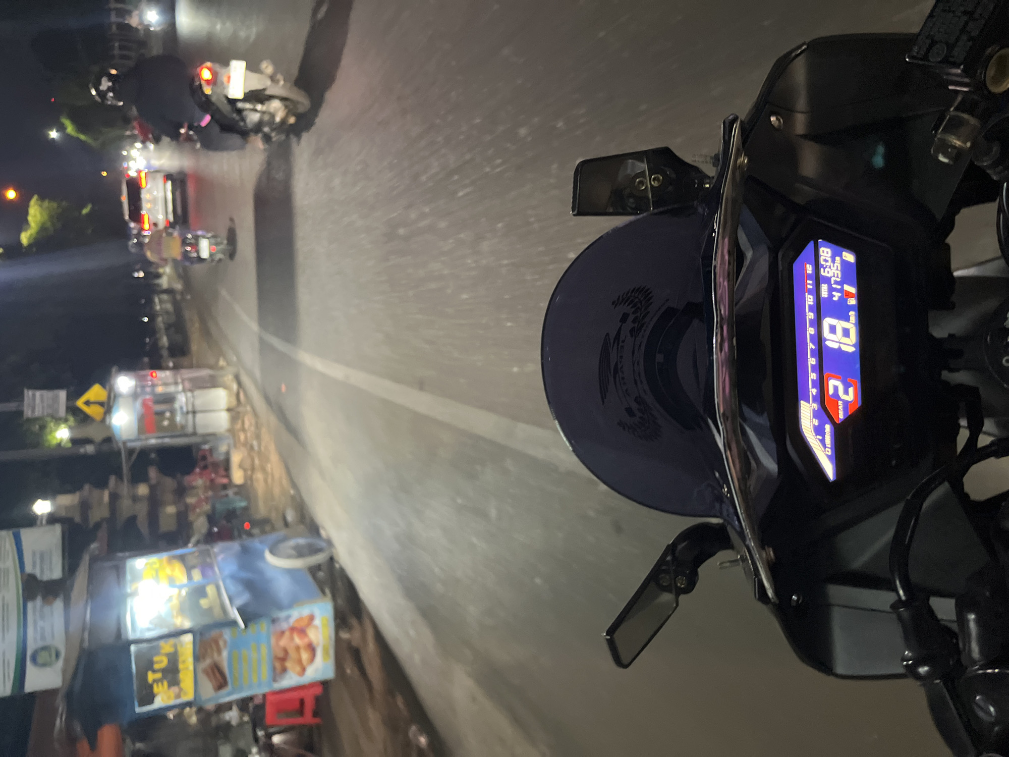 A first-person perspective shot captures a motorcycle's digital dashboard illuminated at night on a road in Pecangaan, Indonesia. The dashboard displays "2" for gear, "18" for speed in km/h, "6:08" for time, and "41735" for mileage, alongside fuel and battery indicators. The rider is proceeding down a paved road, marked with a white dividing line. To the left, a food stall is visible with its goods illuminated. Further down the road, other vehicles, including motorcycles and a white SUV, are also present. The scene is lit by streetlights and the headlights of vehicles, indicating a typical nighttime urban environment.
