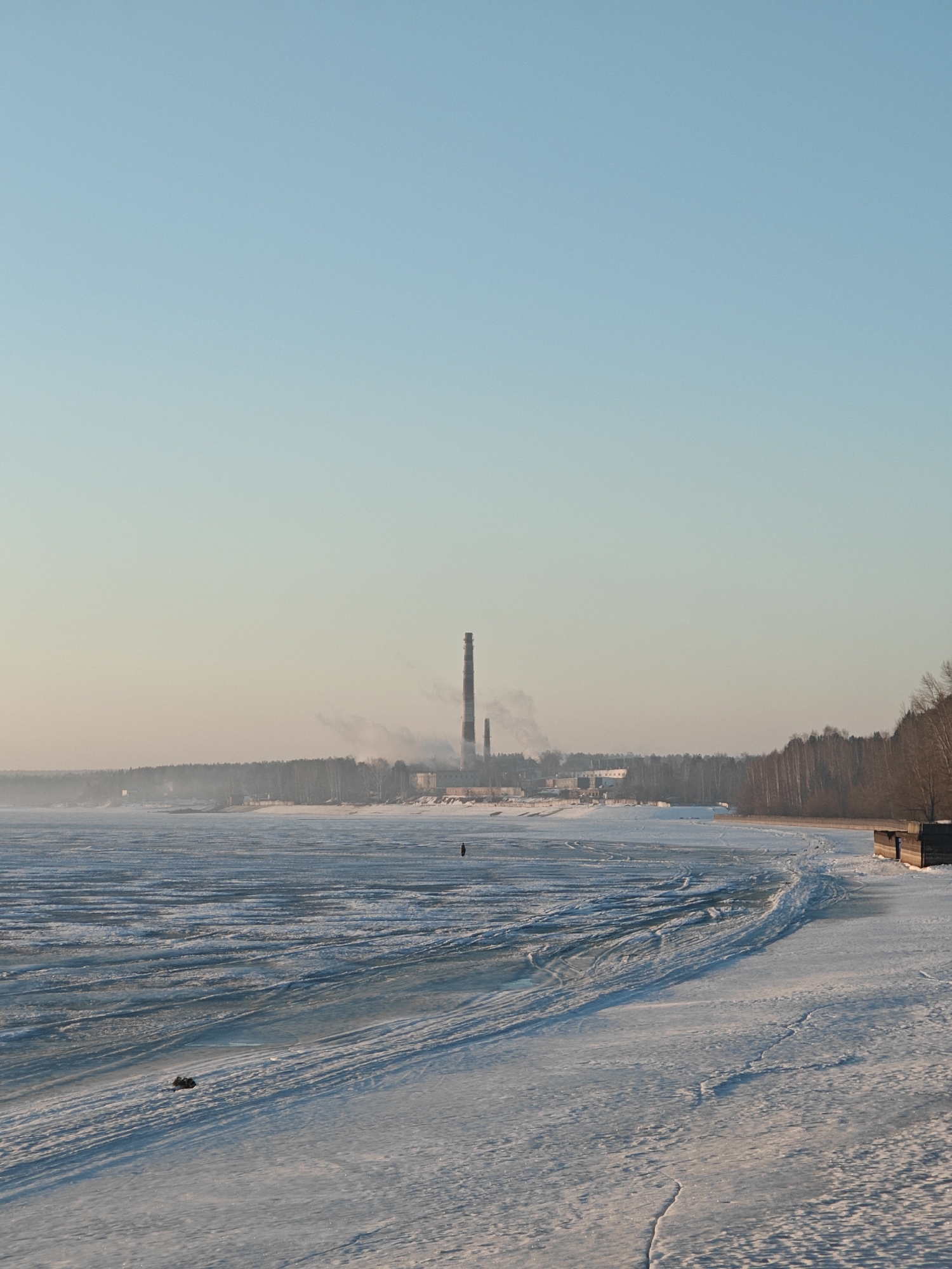 The image depicts a winter scene in what appears to be a coastal area near the city of Chaikovsky, Russia. A partially frozen body of water stretches into the distance, covered in a mix of ice and snow. Tire tracks are visible on the icy surface, indicating vehicular activity.
In the background, a forested area borders the water's edge. Rising above the trees are two industrial smokestacks, suggesting the presence of a factory or power plant. Smoke or steam is emanating from one of the stacks. A lone person can be seen standing on the ice. A small, unidentifiable object is visible on the ice closer to the foreground. Along the right edge of the image is a small wooden structure. The sky is a gradient of light blue, indicating a clear day.