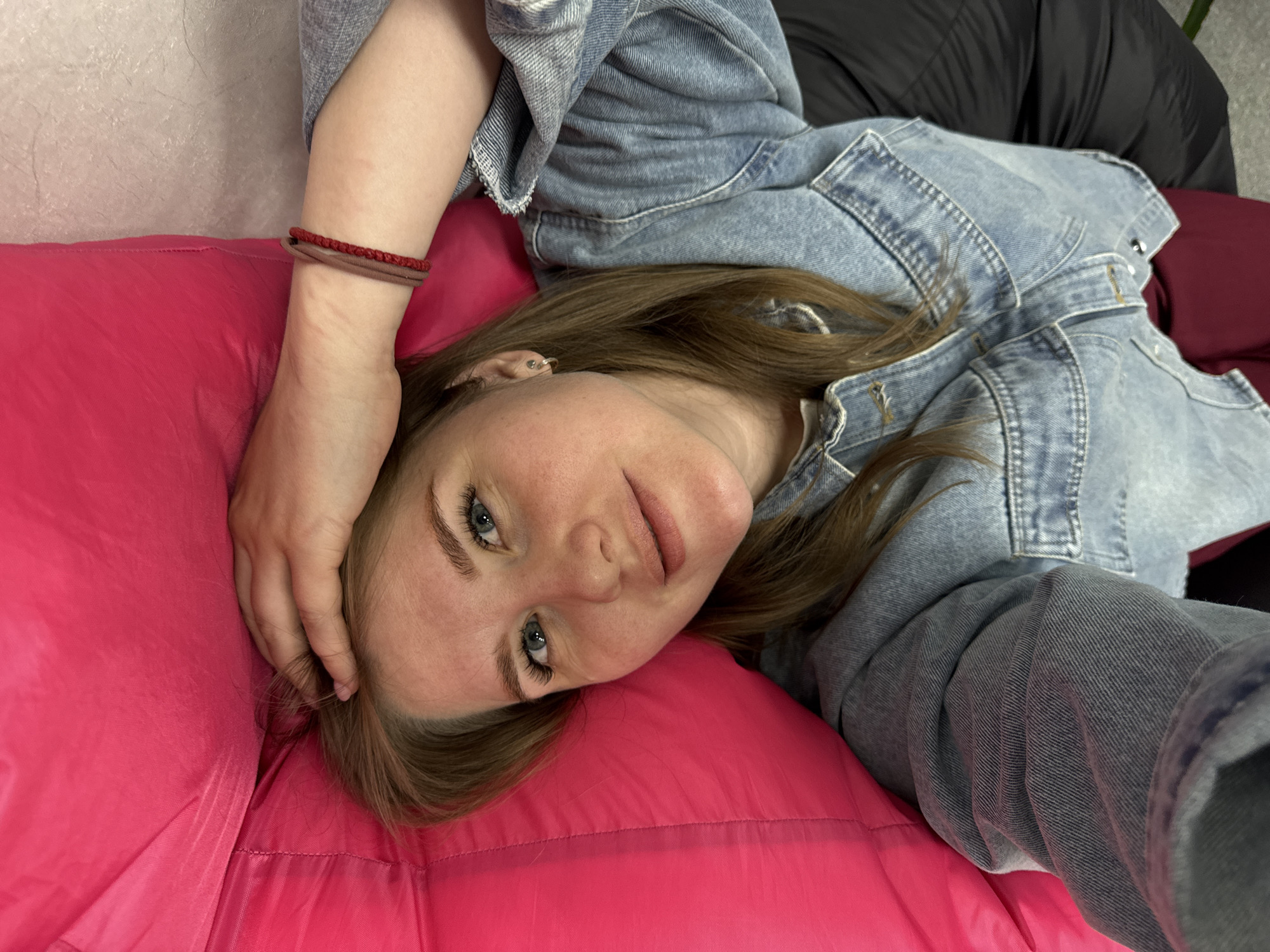 A young woman with light brown hair and blue eyes is lying on a bright pink cushion. She is wearing a light-wash denim jacket. Her right hand rests on her head, and she has a bracelet on her wrist. The background includes a textured white wall and a maroon/black surface. The image suggests a casual, relaxed setting, and the composition appears to be a selfie taken by the woman herself. There is nothing in the image to indicate the location as the city of Perm, Russia.