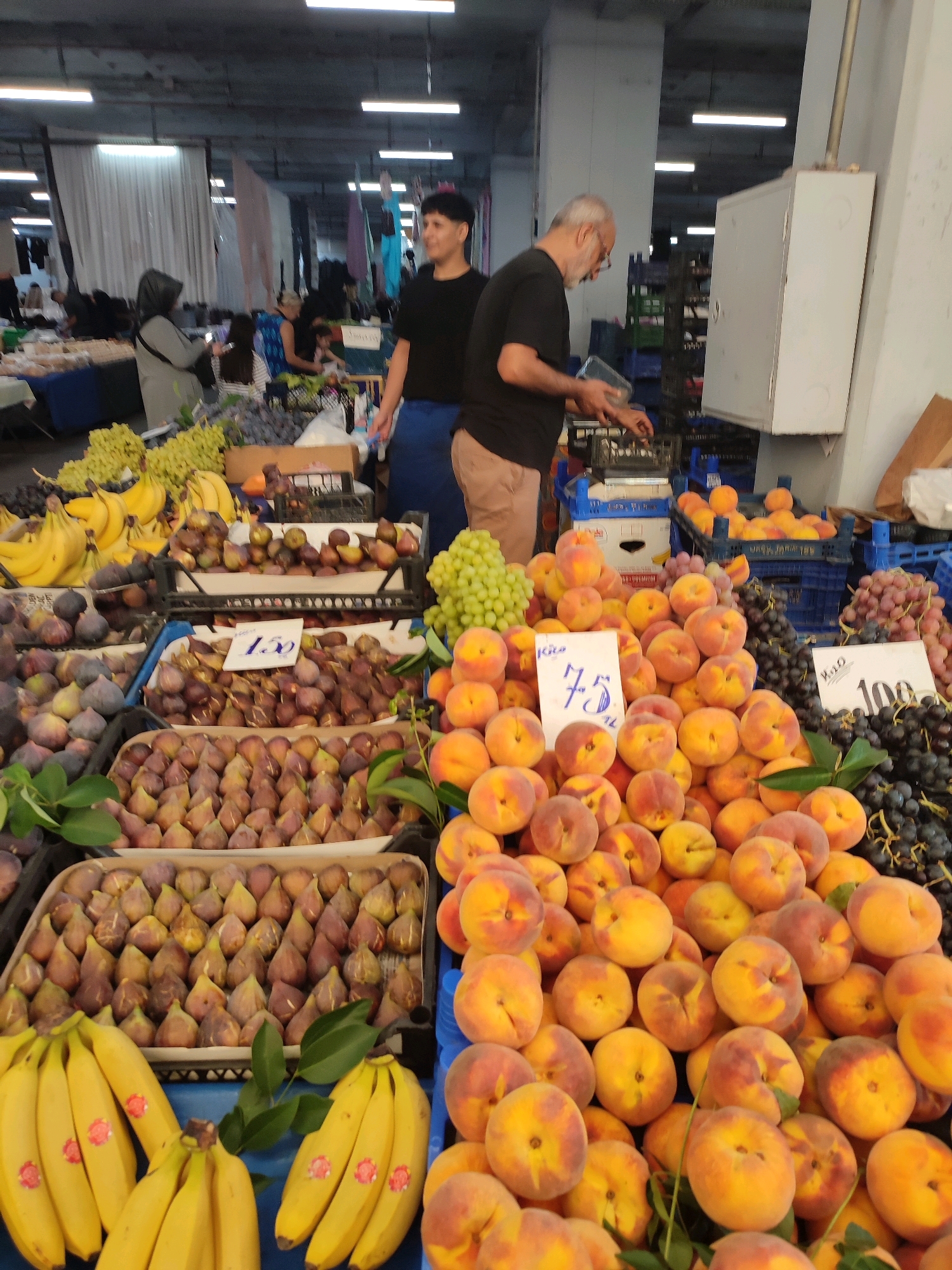 The image depicts a bustling fruit market in Kadıköy, Turkey. Several stalls are visible, laden with a colorful assortment of fresh produce, including bananas, grapes, figs, and peaches. In the foreground, a vendor, likely a man with a beard and glasses, is seen attending to his stall. Further back, another person, a woman wearing a black t-shirt and blue apron, appears to be assisting customers. The market seems to be covered, with a corrugated metal ceiling and fluorescent lighting illuminating the scene. The overall impression is one of a lively and busy marketplace, showcasing the abundance of fresh fruits available in Kadıköy.