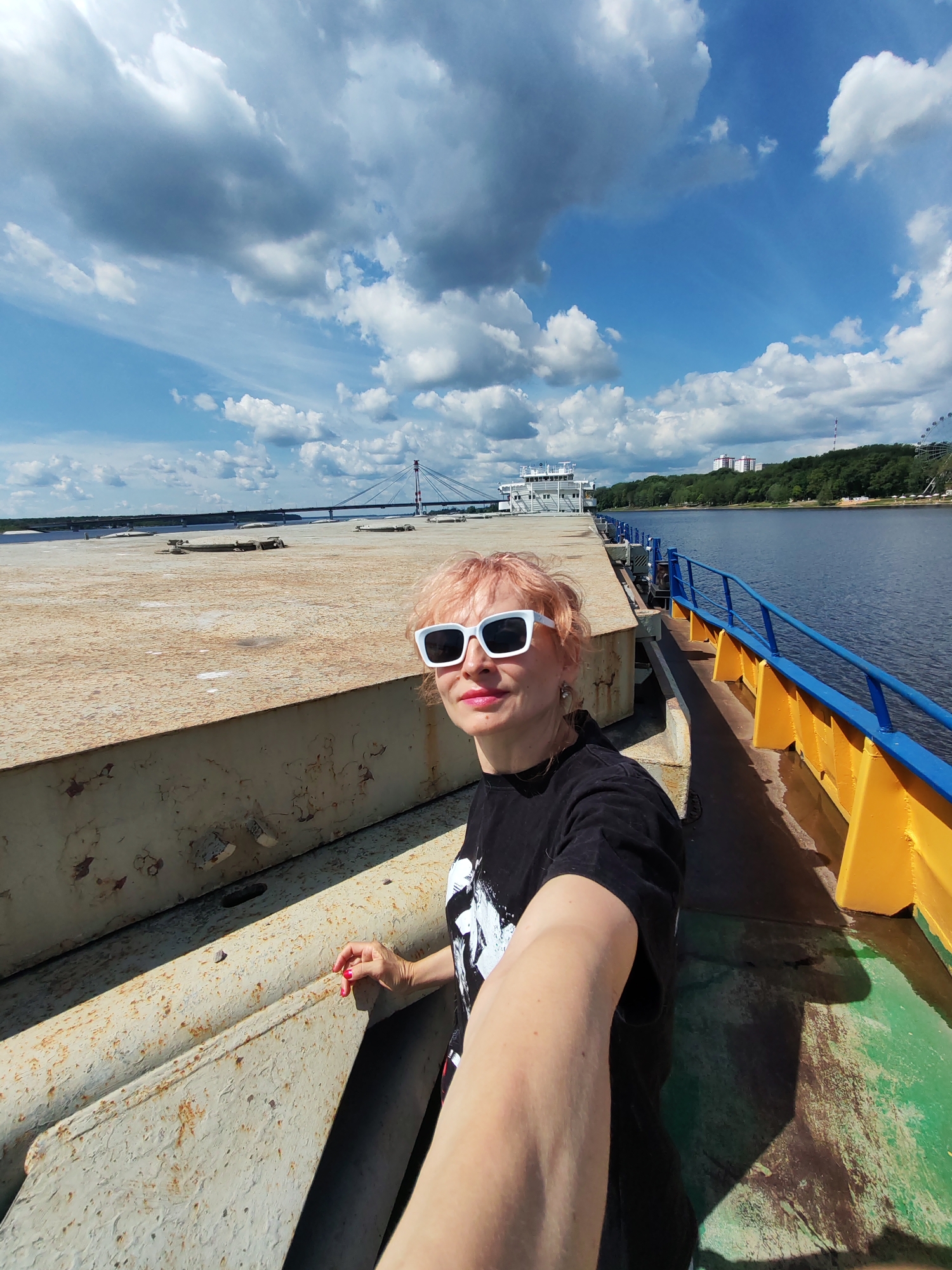 A woman with short, light pink hair and white-framed sunglasses takes a selfie on a large, flat-topped barge. She is wearing a black t-shirt with a white graphic. The barge is made of weathered, rusty metal. To her right, a yellow and blue railing runs along the edge of the barge, overlooking a wide, calm river. In the background, a large white boat is docked, and beyond that, a suspension bridge crosses the river. The scene is set under a bright blue sky with scattered white clouds, suggesting a sunny day. The background also shows a green, tree-lined shore with some buildings and a Ferris wheel. The overall mood appears relaxed and cheerful. The image was taken in Cherepovets, Russia.