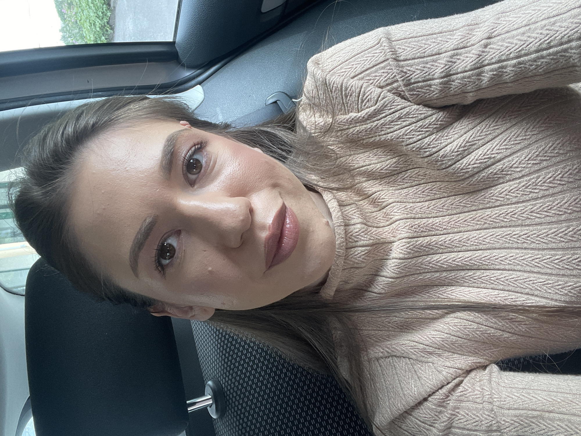 A woman is seated in the driver's seat of a car. She is fair-skinned with light brown hair. She is wearing a light brown, textured sweater. The car interior is dark colored. The location cannot be determined from the image.