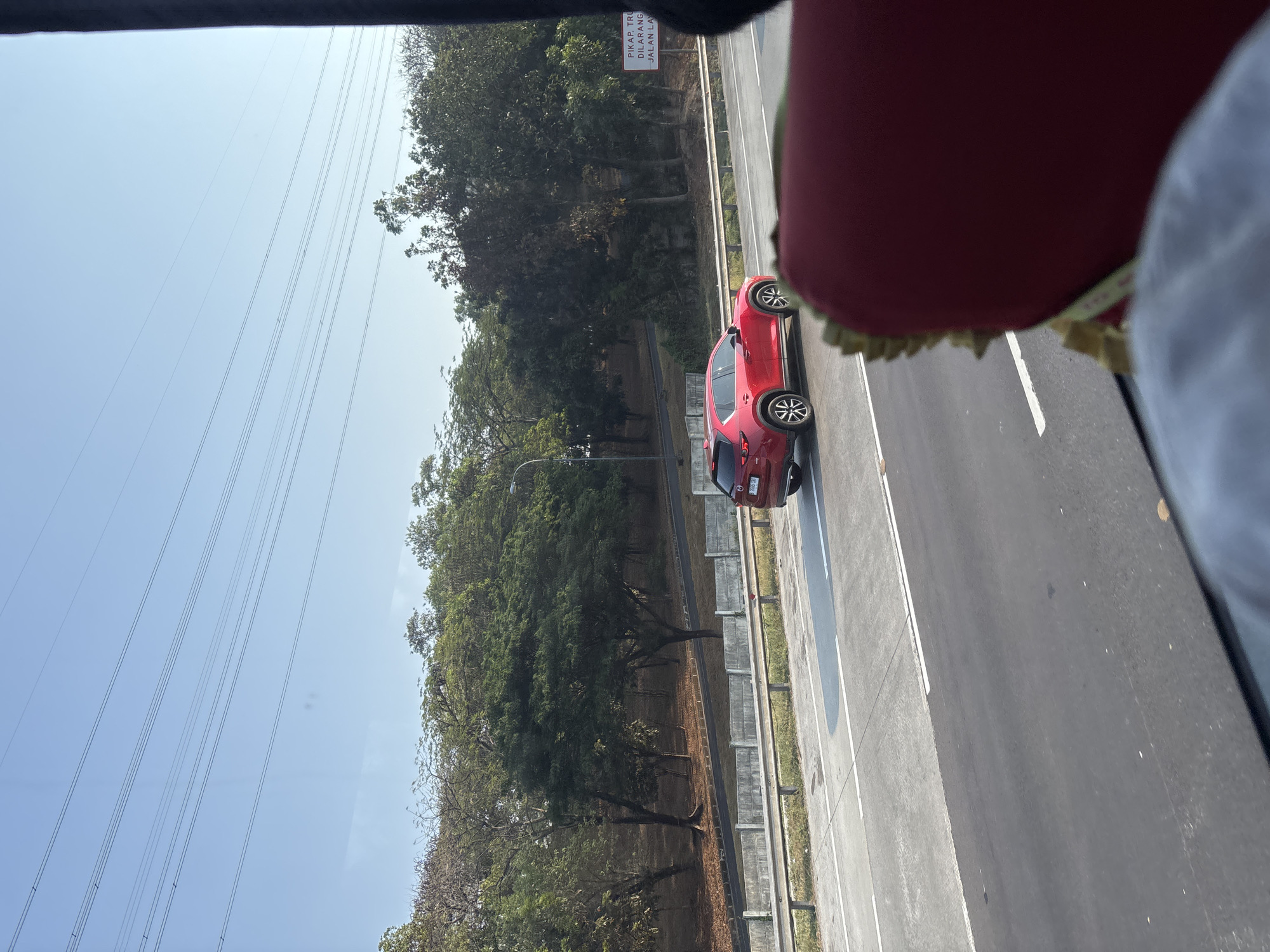 The image captures a clear, sunny day on a highway in Karawang, Indonesia. A red Mazda CX-5 SUV is traveling in the middle lane. The vehicle is seen from the side, with its rear right quarter visible. The car has a license plate that appears to read "B 2023 SAY".
The highway has multiple lanes separated by white lines. To the left of the road, there's a concrete barrier and then a slope with sparse trees and dry leaves on the ground, suggesting a dry season. Further in the background, a dense line of green trees forms a backdrop. Overhead, power lines stretch across the sky.
On the right side of the highway, a white sign is visible, with text that appears to be in Indonesian. Partially obscured, the text seems to include "PIKAP TRU DILARANG JALAN LA". The presence of this sign indicates traffic regulations.
In the foreground, a portion of a red interior, possibly the back of a seat in a vehicle, is visible, suggesting the photo was taken from another vehicle. Part of a white fabric is also visible, likely upholstery. The overall scene depicts a typical highway environment with moderate traffic and the natural landscape of Karawang.
