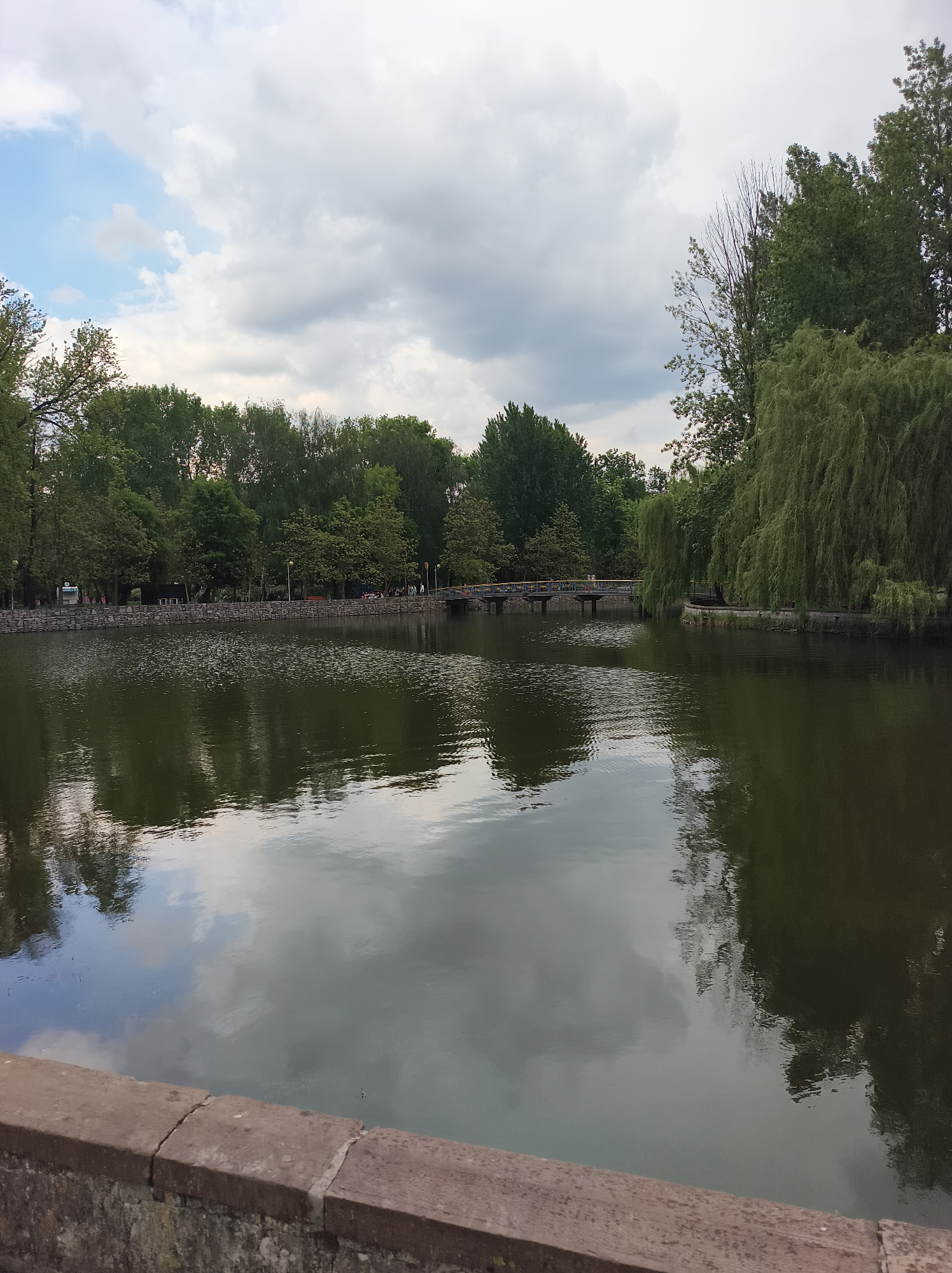 A landscape photograph depicts a lake surrounded by trees in Ternopil, Ukraine. A low wall is visible in the foreground. The lake is relatively still, reflecting the cloudy sky and the surrounding trees. In the middle ground, a small pedestrian bridge crosses the lake. Trees line the perimeter of the lake, including a weeping willow on the right side. People are visible on the far side of the lake. The sky is overcast with patches of blue peeking through.