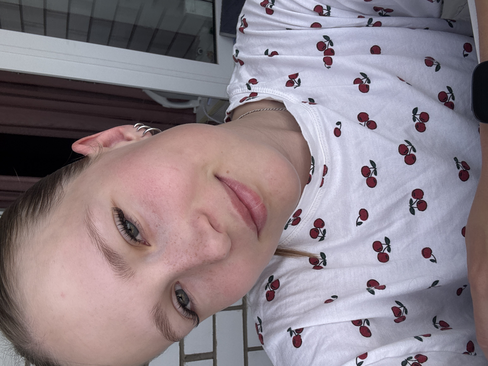 The image shows a close-up, overhead shot of a young woman with light skin, freckles, and grey-blue eyes. She has long, dark eyelashes and her hair is pulled back. She is wearing a white t-shirt with a repeating pattern of red cherries. She has on silver hoop earrings. The background appears to be indoors with a window and curtains visible. The location is not specified in the image or provided crops.