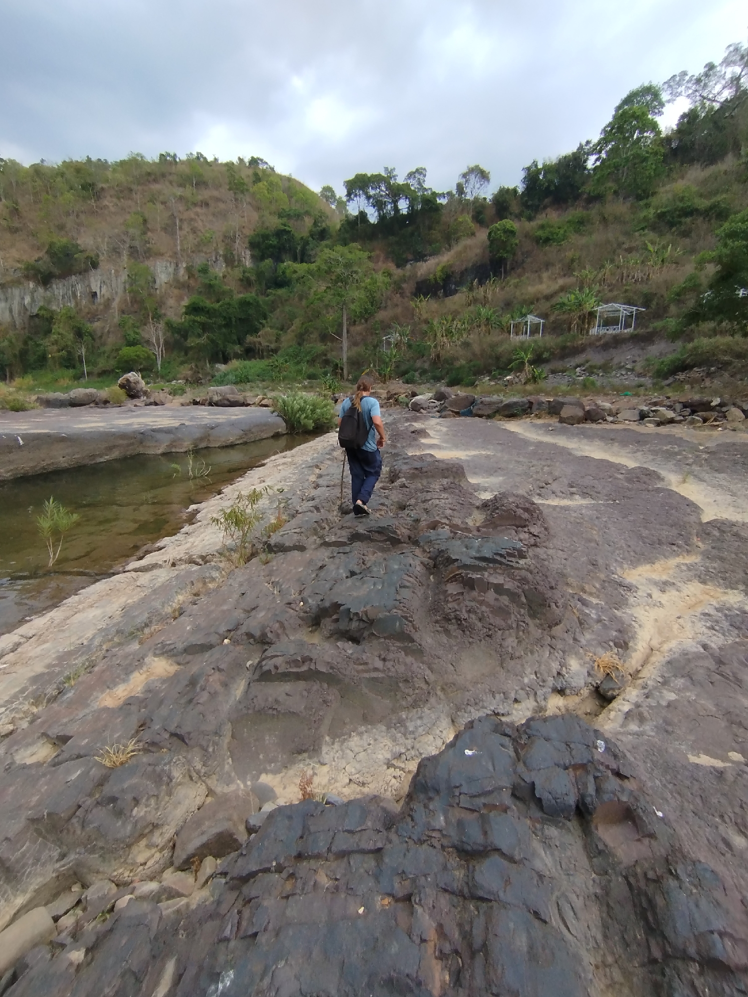 A person wearing a blue shirt and dark pants, with a backpack, is walking away from the viewer across a rocky landscape. The person is holding a stick. To the left, a narrow channel of water separates the rocky area from a smooth, light-colored stone shelf. The landscape rises to a hillside covered in vegetation, with some bare spots. There are small white structures visible on the hillside. The sky is overcast. The location is in the city of Tân Phú, Vietnam.