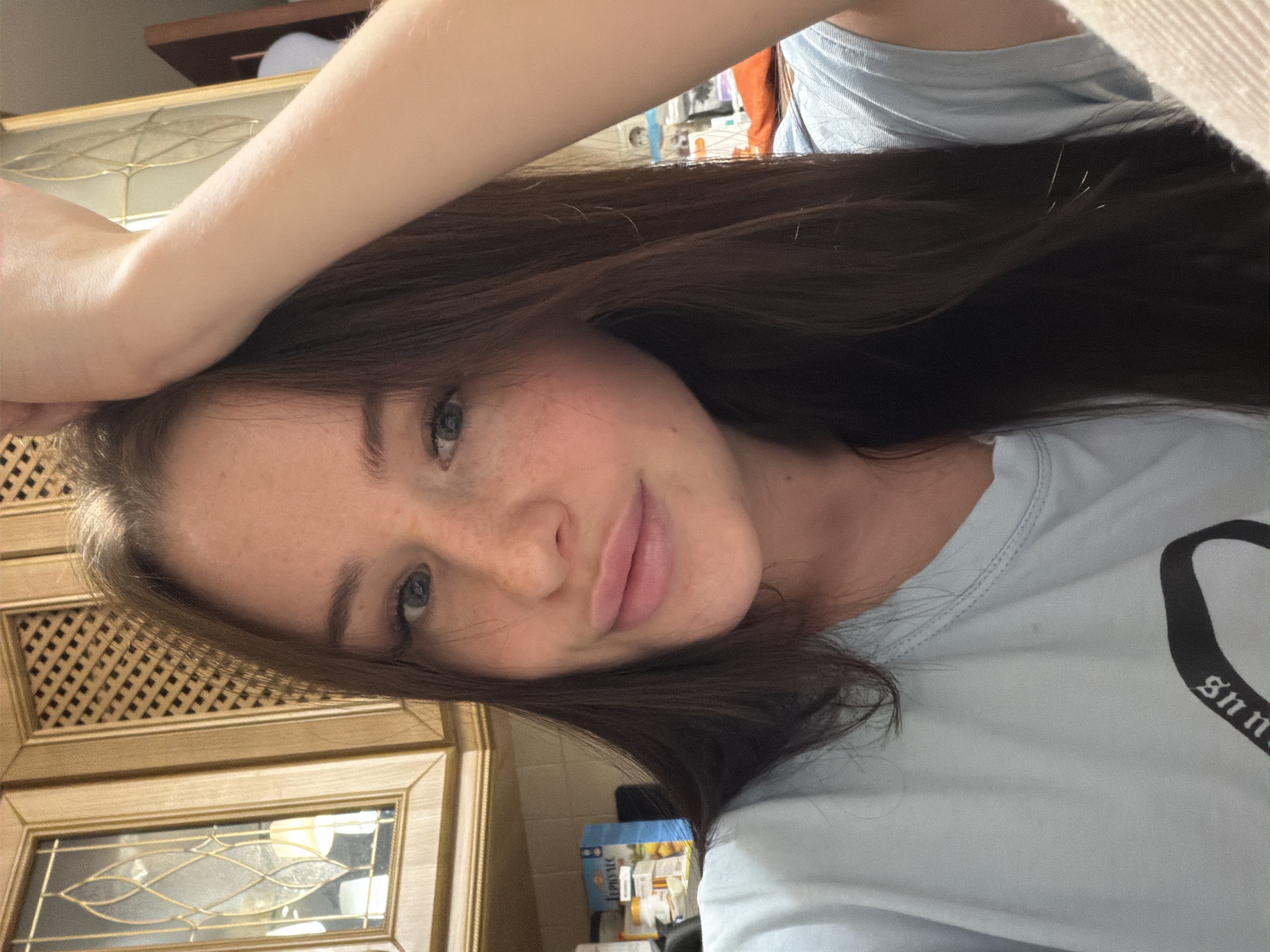 A young woman is depicted in a close-up, rotated selfie. Her long, dark brown hair cascades around her face and across her shoulders. She has blue eyes and a subtle scattering of freckles across her nose and cheeks. Her lips are slightly parted in a gentle, soft smile as she looks directly at the camera. Her left arm is raised and bent, with her forearm resting above her forehead, partially concealed by her hair.
She is wearing a light blue t-shirt. A portion of a dark-colored design or word, possibly in a gothic-style font, is visible on the shirt, appearing to read "gnn" or similar text.
The background suggests an indoor, domestic setting. To the left side of the image (corresponding to the right of her head in a correctly oriented view), there are light-colored wooden cabinets or furniture with decorative lattice patterns and glass panels. To the right, a blurred area reveals what appears to be a counter or shelf holding various household items, including a blue box. The lighting is soft and natural, indicating it is likely daytime.