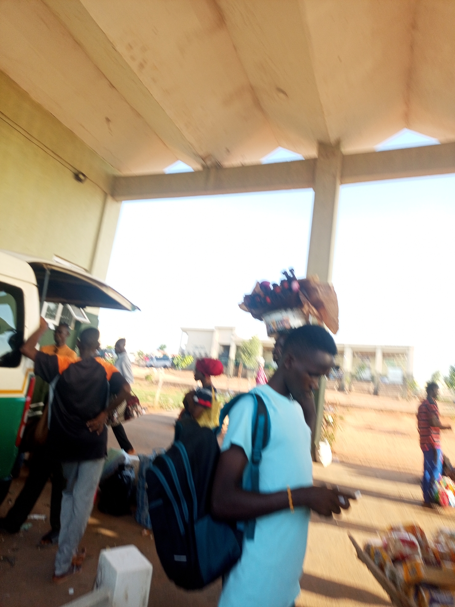 The image shows a group of people at an outdoor transit station in the city of Unknown, Unknown. Several individuals are near a white van with a green stripe, possibly loading or unloading luggage. One man is wearing a light blue t-shirt with a dark backpack, and another man carries goods on his head. A cart filled with packaged food is in the foreground. The location appears to be a transportation hub or terminal.