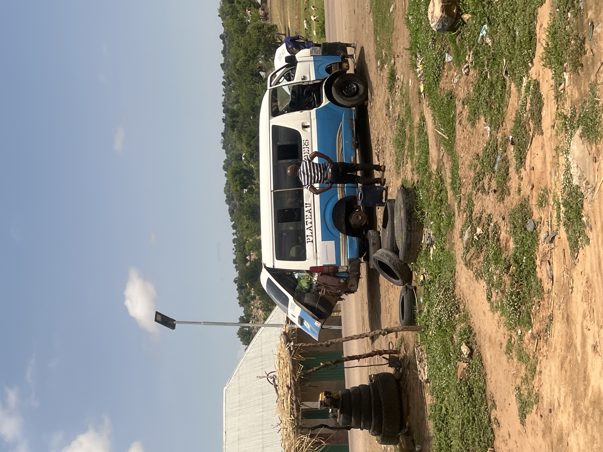 The image displays an outdoor scene in Sabon Layi Jessu, Nigeria, under a bright blue sky with scattered white cumulus clouds. A white and blue passenger van, bearing the partial inscription "PLATEAU DERS" on its side, is central, appearing to be undergoing roadside repair. The van is missing its rear driver-side wheel, and the front passenger-side wheel is also either removed or being worked on. Its front passenger door is open, and the rear hatchback is ajar, revealing interior cargo and spare tires.
Two men are visible near the van. One man, dressed in a black and white striped short-sleeved shirt and dark pants, stands with his back mostly to the camera, observing the van's side. A small blue rolling luggage bag rests near his feet. The second man, wearing blue attire, is partially obscured on the passenger side of the van, seemingly engaged with the front wheel area.
To the left of the van, a rudimentary structure with a thatched roof supported by wooden poles stands on a dirt patch. Beneath the shelter, a tall stack of discarded black tires forms a conical shape, topped by a yellow portable generator or engine. A bicycle frame is visible on the roof of the shelter. The ground around the van and shelter is predominantly brown dirt, interspersed with patches of green low-growing vegetation and some scattered litter, including plastic debris and wrappers. Several individual black tires are also strewn across the ground.
A modern solar-powered street light with a dark, rectangular panel is positioned on a silver pole behind the makeshift shelter. In the middle ground, behind the van, a paved road transition gives way to a dirt track. The background features lush green trees covering undulating hills, with a few distant houses or structures and several grazing animals, possibly goats or sheep, discernible on the right side. A building with a silver corrugated metal roof is visible behind the shelter.