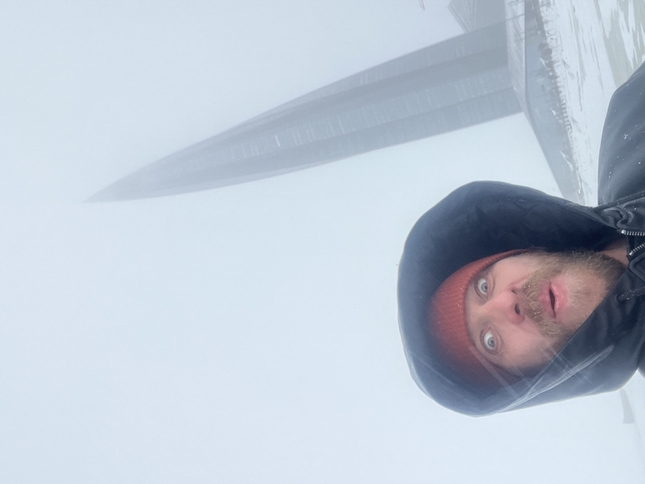 An individual with a beard and mustache, wearing a rust-colored knit beanie and a black hooded jacket, is depicted in a close-up, selfie-style photograph. The person exhibits a wide-eyed, open-mouthed expression, suggestive of surprise or humor. Snowflakes are visible on their beard and jacket.
In the background, positioned in the upper right, the Lakhta Center skyscraper is partially visible, its distinctive tapering structure piercing through dense fog or low clouds that obscure its upper sections and the surrounding sky. Below the skyscraper, a lower building with a dark, sloping roof and glass facade, likely part of the Lakhta Center complex, is also partially discernible. The ground in the mid-ground and foreground is covered in snow and ice, indicating winter conditions. The sky is uniformly white-gray due to the heavy atmospheric obscuration. The scene is set in Saint Petersburg, Russia.