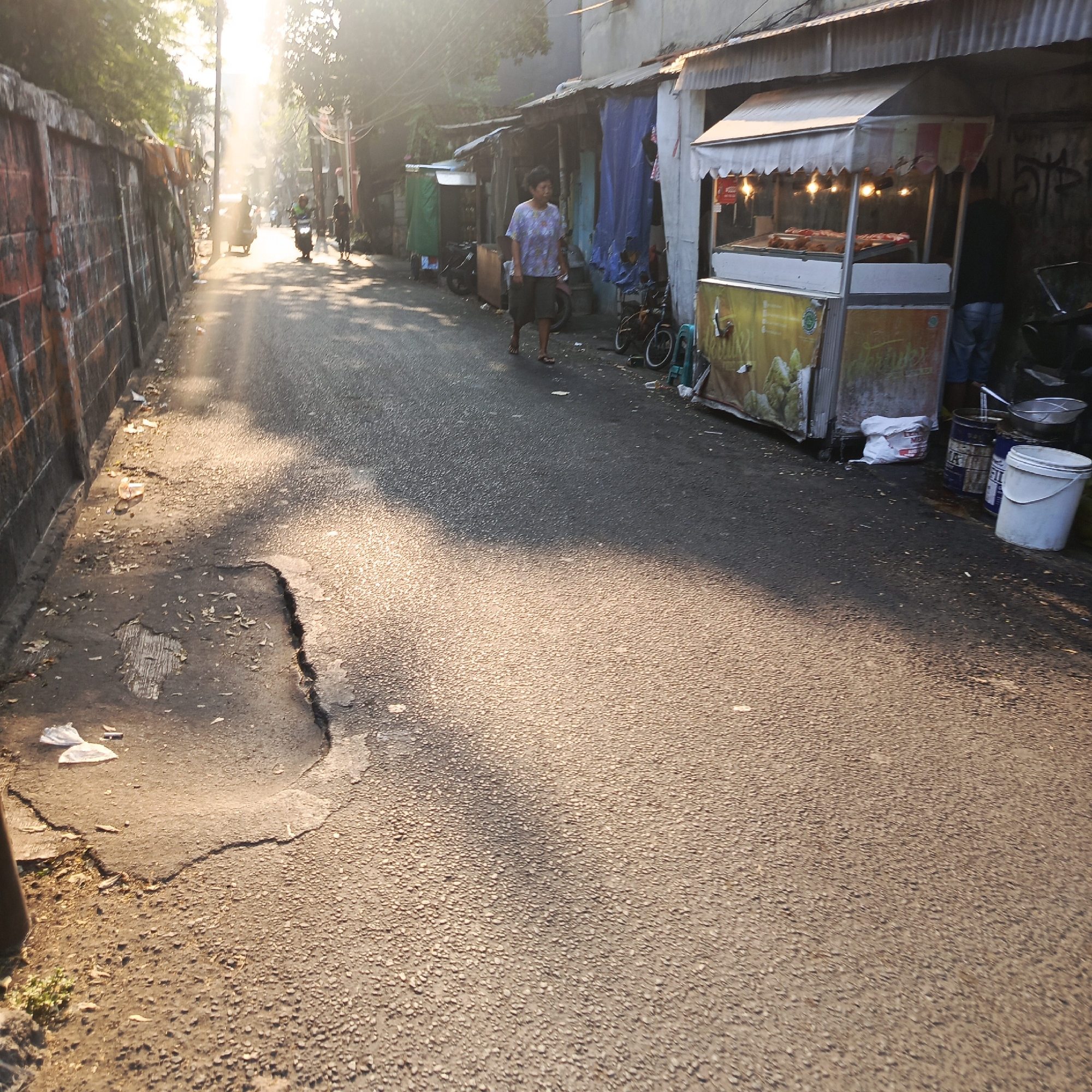 The image shows a sunlit street scene in South Jakarta, Indonesia. A man walks down the street, passing food stalls and various shops lining the road. The street is paved and shows signs of wear and tear, with a large pothole visible on the left side. Sunlight streams through trees, casting long shadows across the road. The stalls appear to be selling food and other items, with goods displayed for sale. A motorcycle is parked near one of the stalls. The overall impression is a bustling, everyday scene in a densely populated urban area.