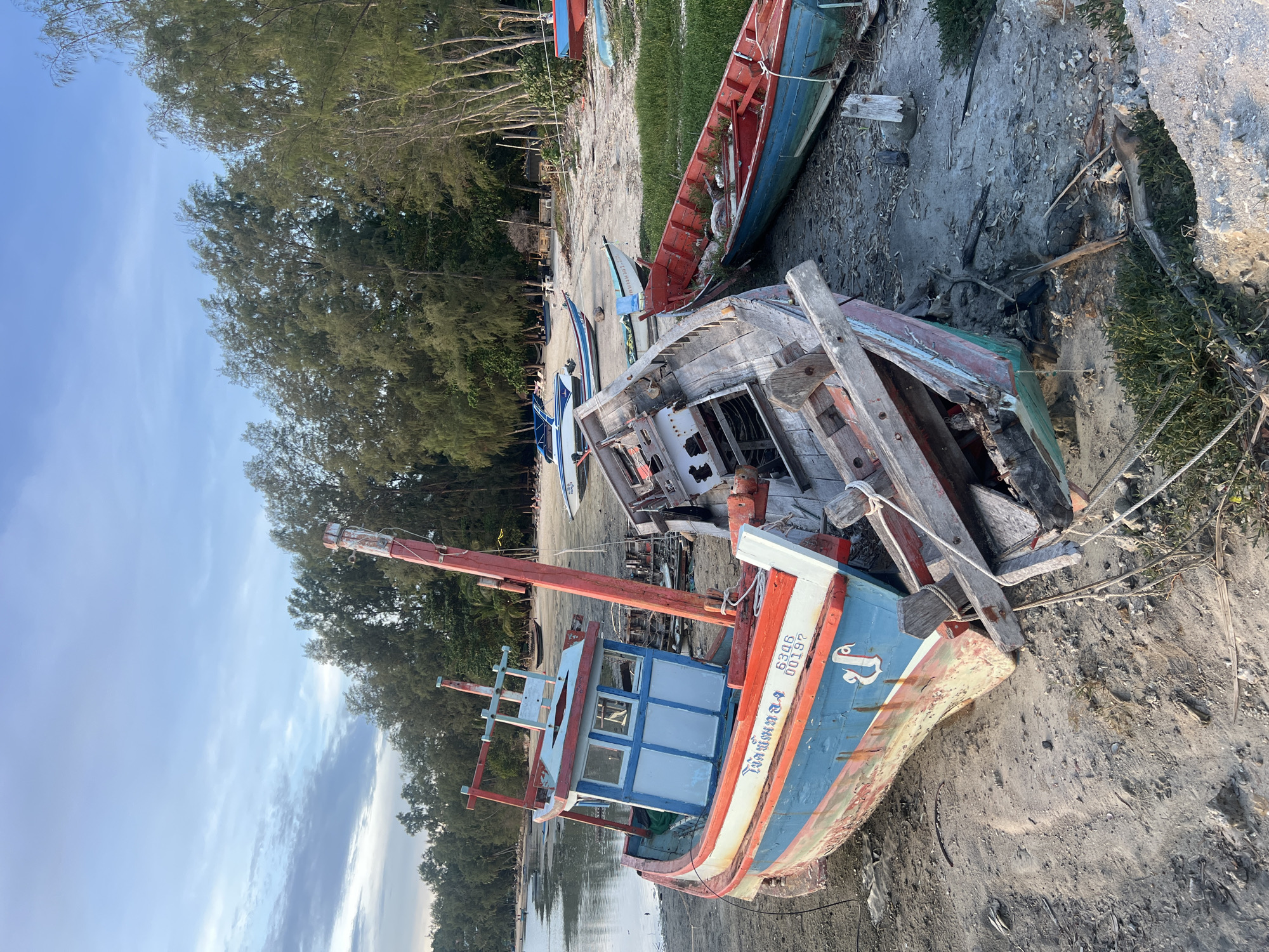 A wide shot shows a beach with several boats, some in various states of disrepair. The main focus is a blue, red, and white fishing boat beached on the sand. It has a wooden cabin with windows and a tall red mast. Another severely damaged wooden boat is partially visible next to it. Further down the beach, more boats are scattered, including a red and green boat and a white speed boat. The background features a line of trees and a calm body of water. The sky is mostly clear with some light clouds.