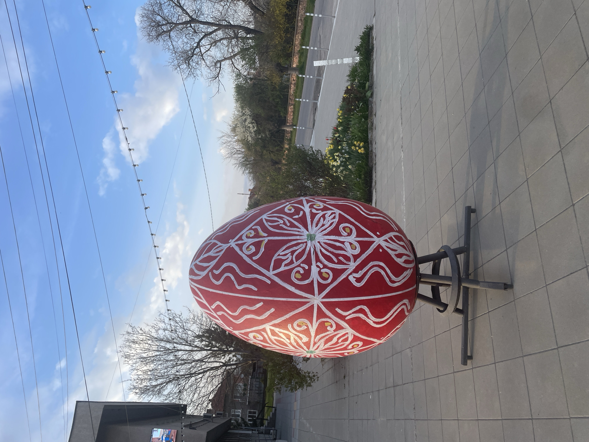 The image shows a large, red, decorated Easter egg on a metal stand in what appears to be a public space. The egg is decorated with white and gold colored patterns. The background includes a road, trees, and buildings, indicating an urban setting. The sky is partly cloudy with power lines visible. The location is Aprilovo, Bulgaria. The camera angle is tilted.