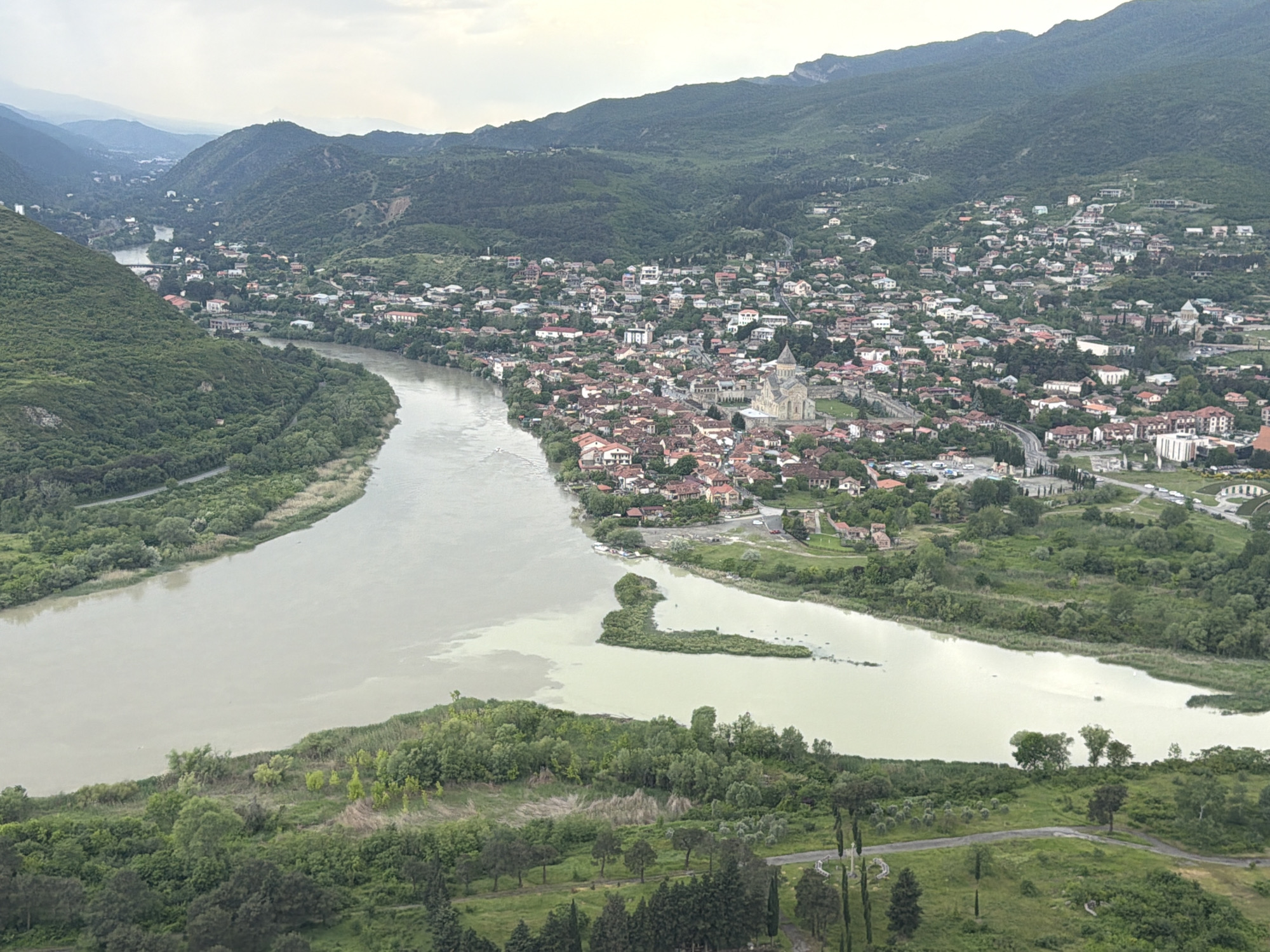 The image presents an elevated, wide-angle view of the city of Mtskheta, Georgia, situated at the confluence of two rivers, the Aragvi and the Mtkvari (Kura). The rivers appear cloudy, with varying shades of brown and tan, reflecting overcast skies. Dense vegetation lines both riverbanks.
The city of Mtskheta is characterized by clusters of buildings, largely residential, with red-tiled roofs. Dominating the skyline is a large, light-colored cathedral complex, presumably Svetitskhoveli Cathedral, near the river junction. Hills and mountains form the backdrop of the city, covered with dense green forests and sporadic settlements. Roads and other infrastructure are visible among the urban and rural landscape. The overall impression is that of a historic, densely populated urban area nested within a valley surrounded by undulating, verdant terrain. The foreground shows a grassy landscape with trees.