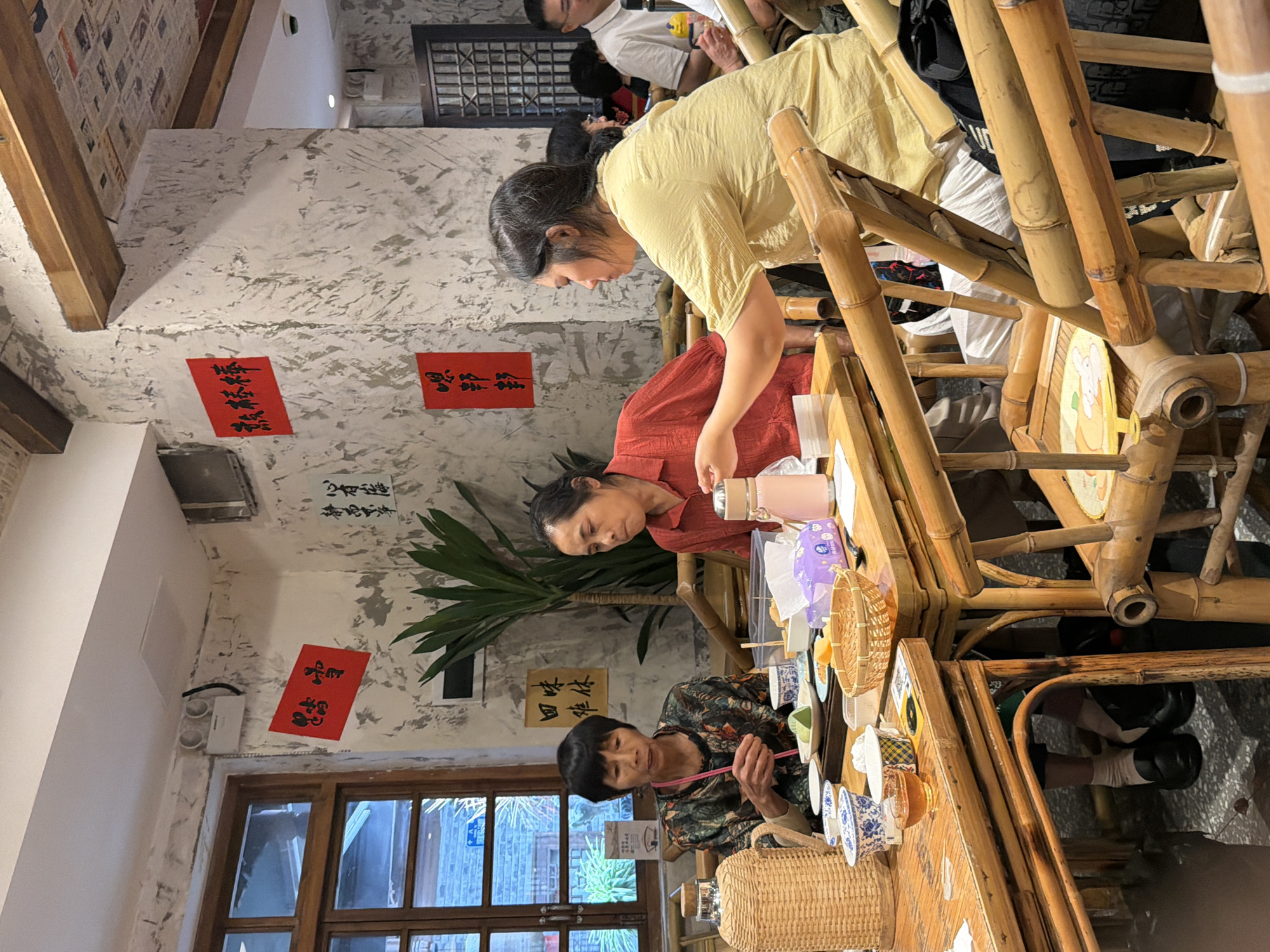 A medium shot captures a group of people at tables in a restaurant in Yuzhong District, China. The tables and chairs are made of bamboo. On the wall, red calligraphy banners are displayed. The atmosphere appears to be casual dining. The focus is on three women and other patrons in the background.
In the foreground, two women are seated at a table. The woman on the left, with short black hair, wears a patterned shirt and is holding a small stick over a plate of green food. The woman to her right, wearing a red shirt, looks down at the table. A third woman, wearing a yellow shirt, is seated further to the right, leaning forward and reaching for something on the table.
On the table, there are various items including bowls, a glass of amber liquid, a woven basket, and a pink thermos. To the left, a large woven water bottle sits on the table. The background shows more tables, diners, and the interior architecture of the restaurant, including a window with a view of greenery and a patterned ceiling.