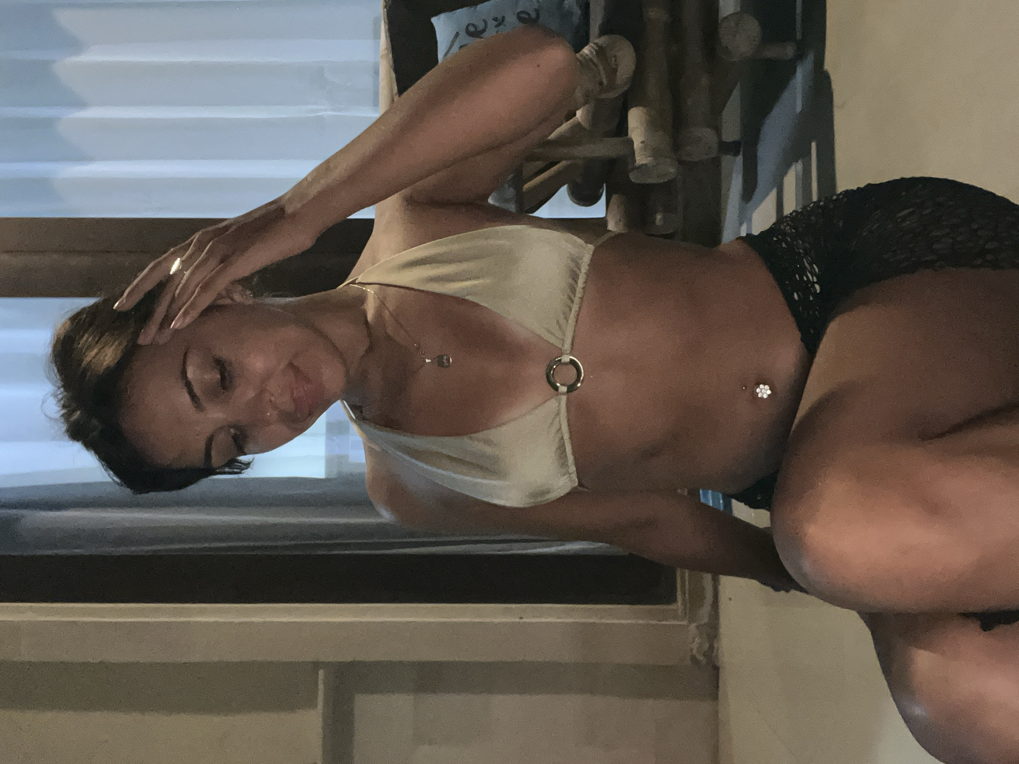 A woman is posing indoors. She is wearing a bikini top and shorts. She has one hand on her head and her hair is pulled back. There is a window behind her and what appears to be a bamboo chair. This may be in Kuta Selatan, Indonesia.