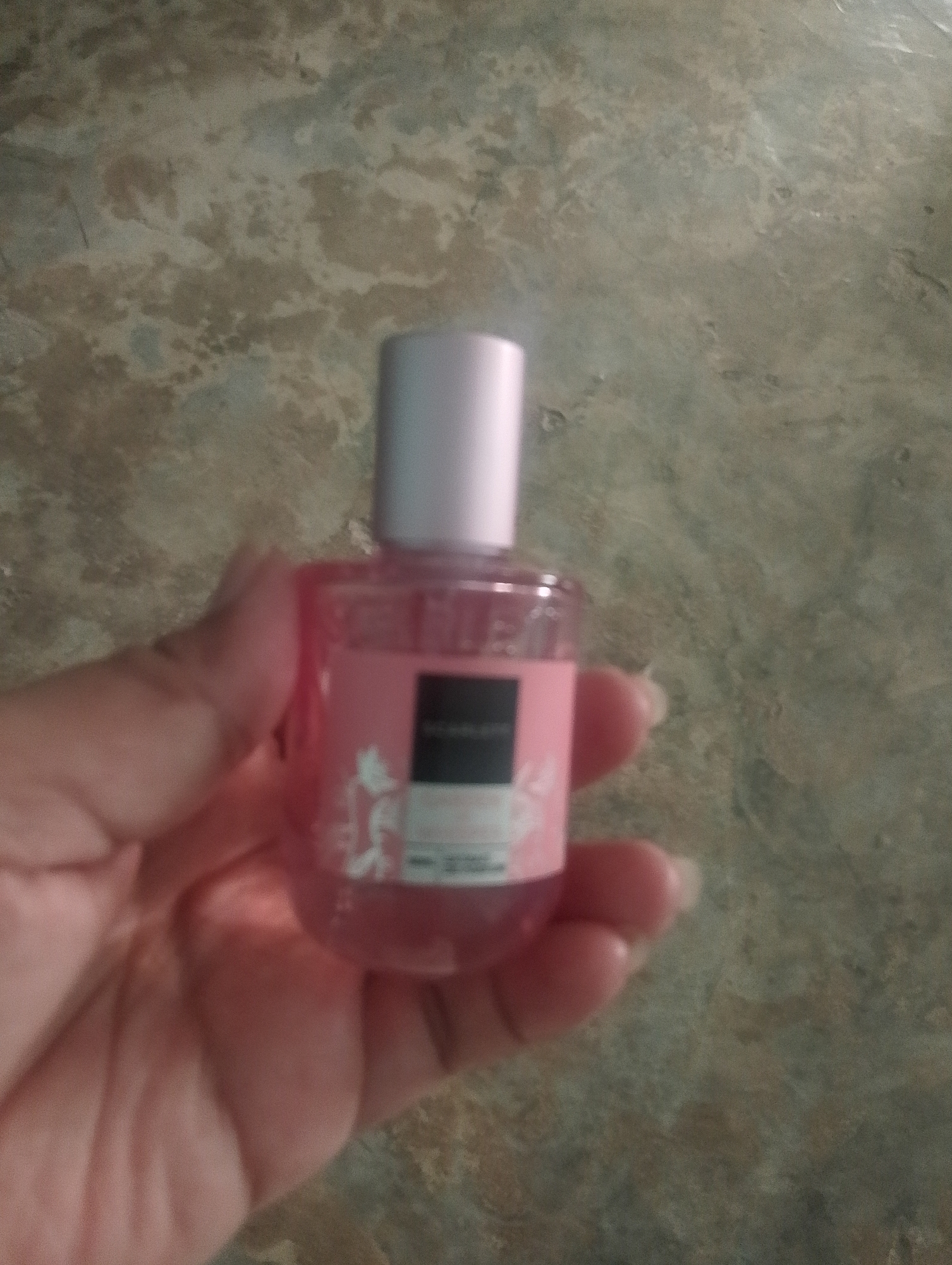A hand holds a small, pink, square-shaped bottle of perfume. The bottle has a silver cap and a label with black text. The background is a textured, grey-brown surface, possibly a floor or wall, with an uneven pattern. The image appears to be taken indoors, with no clear indication of Serbelawan, Indonesia.