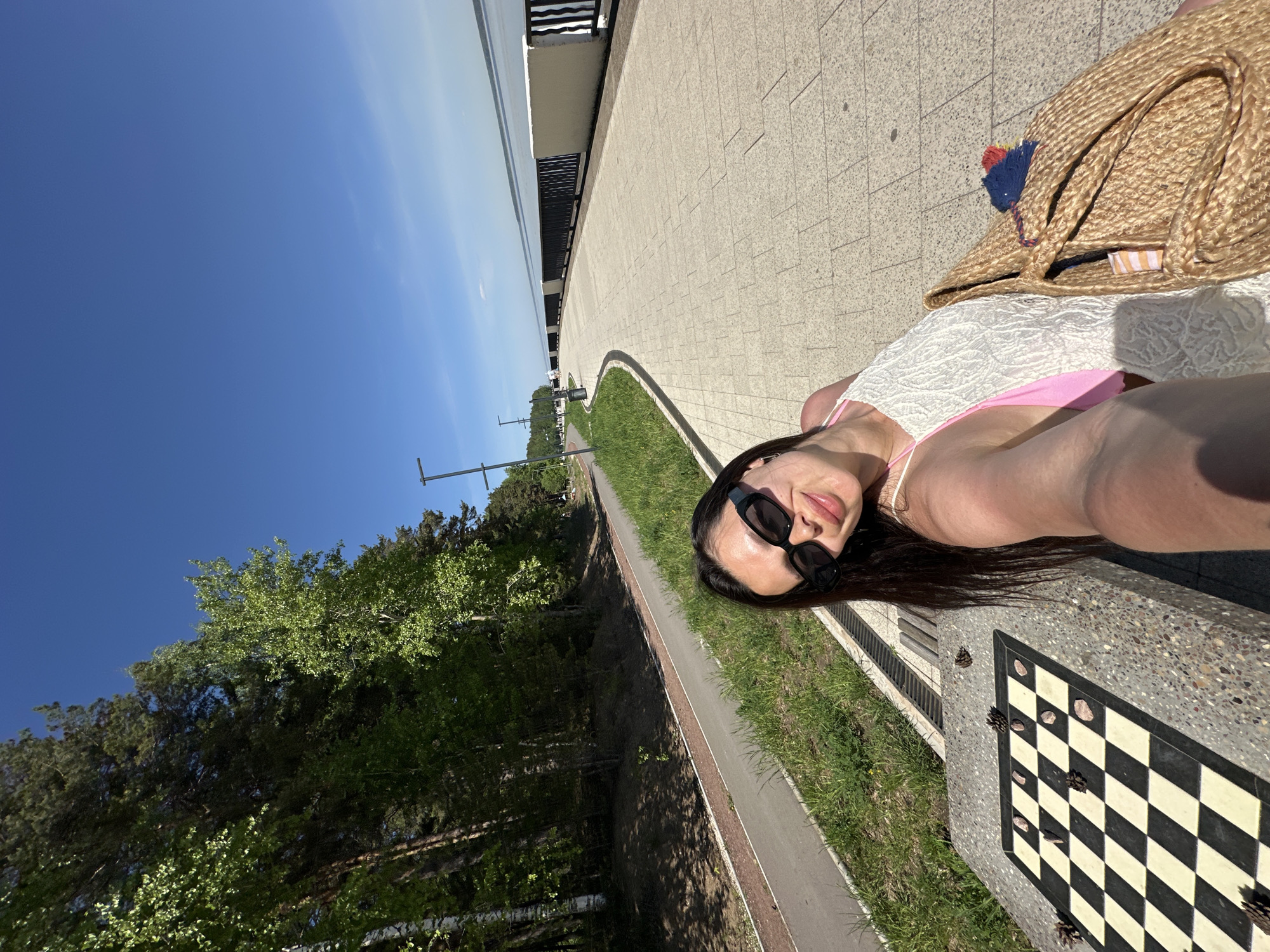 A woman is taking a selfie while lying on a bench next to a chessboard table. She is wearing sunglasses, a white cover-up, and a pink bathing suit. A straw tote bag is beside her. The scene is outdoors, along a paved walkway with grass and trees on one side and a body of water in the distance. Based on the chessboard table, the image is likely taken in the city of Chaikovsky, Russia.