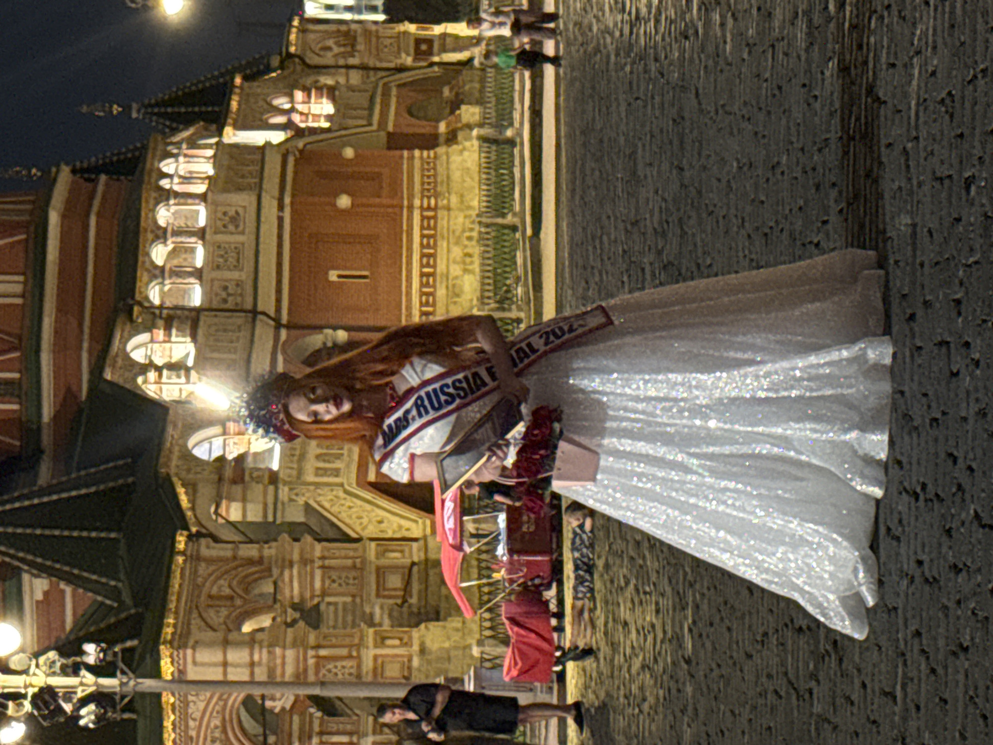A woman with long auburn hair and a crown stands in the center of the image, dressed in a sparkling white gown and a sash that reads "MRS. RUSSIA FESTIVAL 2023". She holds a framed photograph and a bouquet of red roses in a small pink purse. Behind her, the iconic red brick buildings and onion domes of St. Basil's Cathedral on Red Square are illuminated at night. Several people are visible in the background, including a man in a black shirt standing to the left, and two children and an adult woman walking further away. A red tented cart, possibly a souvenir stand, is also present. The ground is paved with cobblestones, reflecting the ambient light.