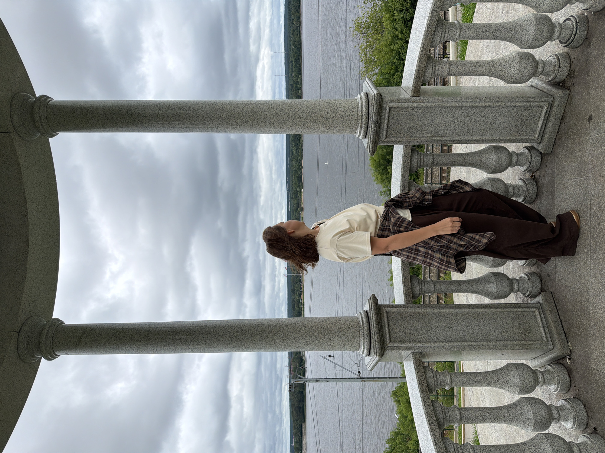 A woman stands on a stone balcony with a railing, looking out at a wide river. She is wearing a light-colored t-shirt, brown wide-leg pants, and a plaid shirt tied around her waist. The balcony is supported by decorative stone columns. In the background, across the river, is a forested shoreline with buildings and what appear to be power lines. The sky is overcast with clouds. The location is the Permskaya Ramena observation deck in the city of Perm, Russia.