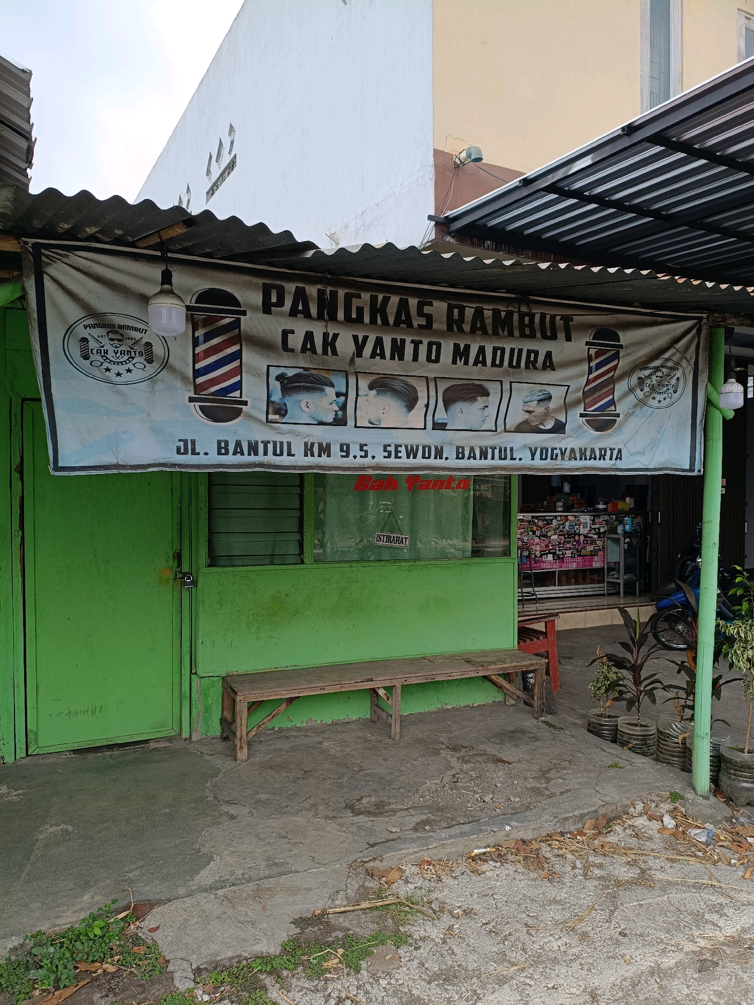 A light blue banner advertising "PANGKAS RAMBUT CAK YANTO MADURA" hangs above the entrance to a barbershop. The banner features images of haircuts, barber poles, and the shop's logo. Below the banner, the address "JL. BANTUL KM 9.5, SEWON, BANTUL, YOGYAKARTA" is visible. The barbershop itself has a green painted door and a green painted facade. A wooden bench sits in front of the shop. The ground in front of the shop is paved with concrete. In the background, a motorcycle is parked. The scene appears to be an outdoor street view in Sewon, Indonesia.