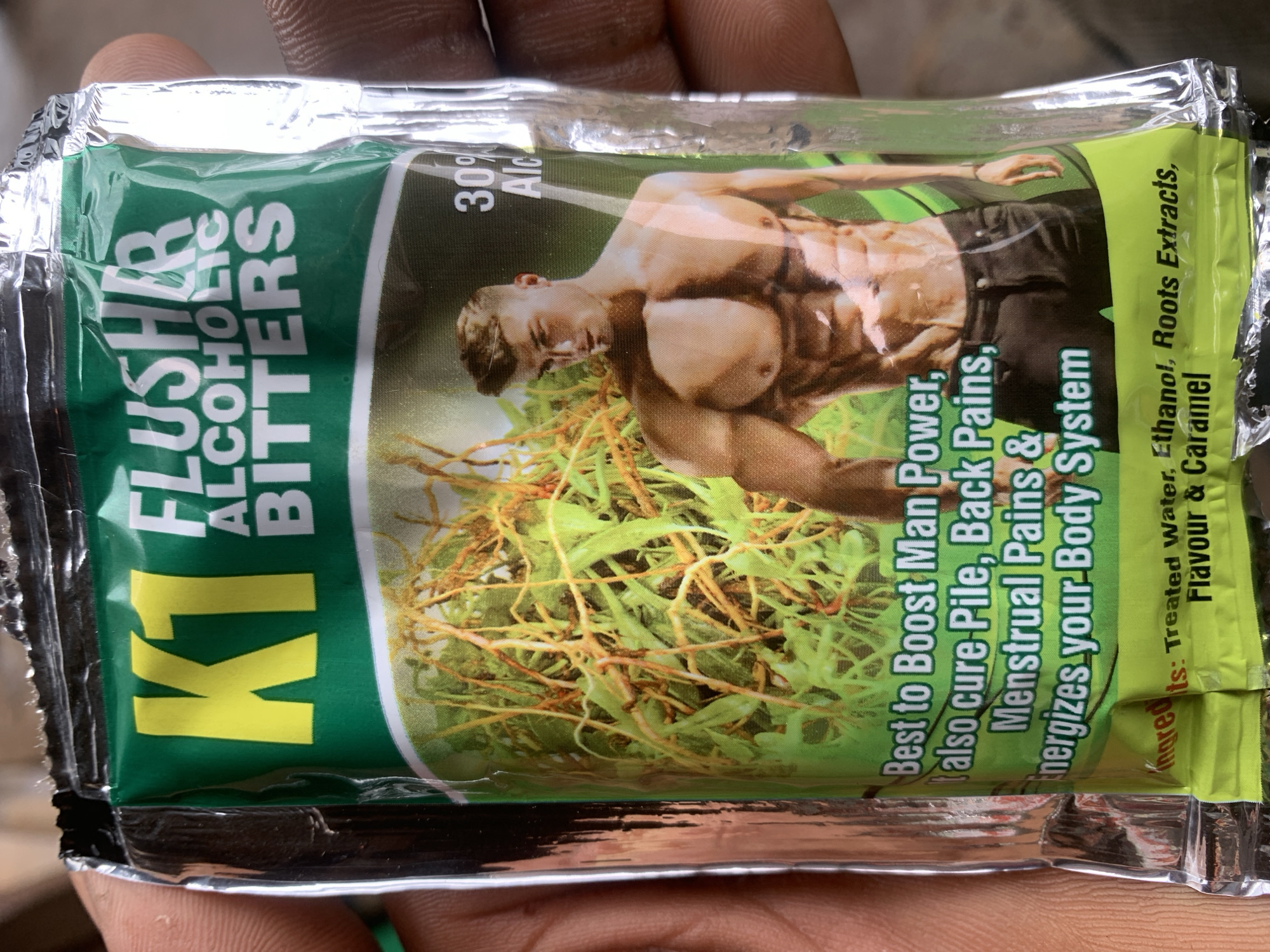 A person's hands hold a sealed packet of "K1 FLUSHER ALCOHOLIC BITTERS" in Shagamu, Nigeria. The packet features a vibrant green background with yellow lettering for "K1" and white lettering for the product name. A stylized image of a muscular man adorns the front, alongside an illustration of roots and green foliage. Text on the packet indicates it is "Best to Boost Man Power, also cure Pile, Back Pains, Menstrual Pains & energizes your Body System." Below this, a list of ingredients includes "Treated Water, Ethanol, Roots Extracts, Flavour & Caramel." The lighting suggests an outdoor or well-lit indoor environment.