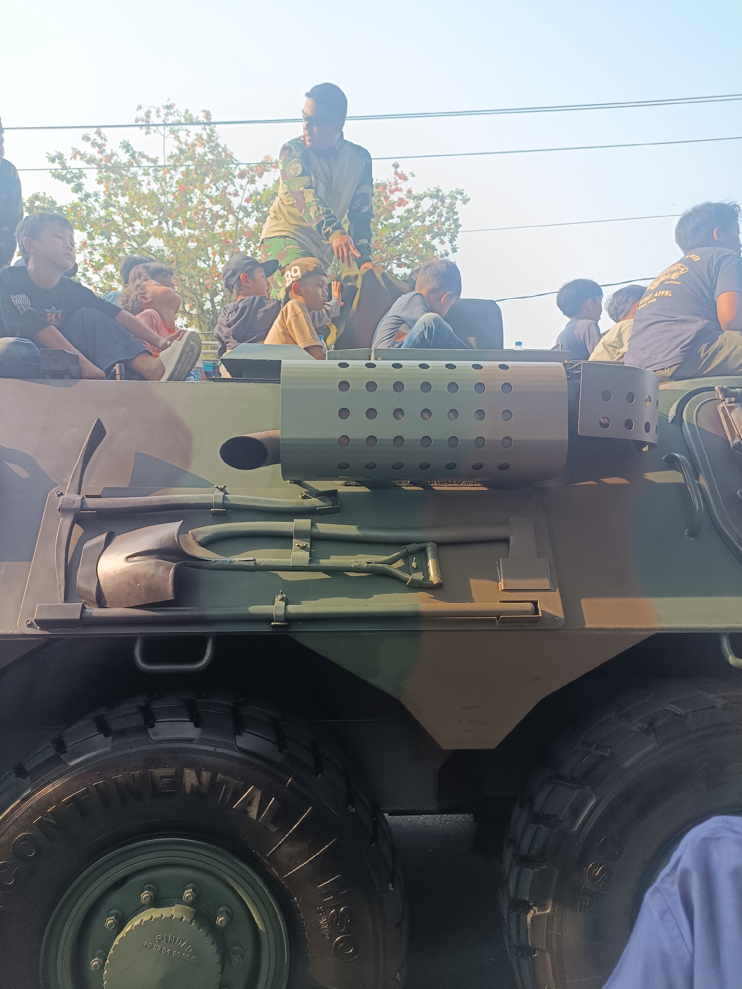 In Sleman, Indonesia, a military vehicle, likely an armored personnel carrier, is parked outdoors, with its large tires and camouflage paint visible. Several children and a soldier are positioned on top of the vehicle, with the soldier appearing to interact with the children. The children are looking up at the soldier, some with expressions of curiosity. The background shows trees and a bright sky, suggesting it is daytime. The vehicle is equipped with tools, including a shovel and a pickaxe, attached to its side. The scene appears to be part of a public event or demonstration, given the presence of onlookers and the soldier's interaction with the children.