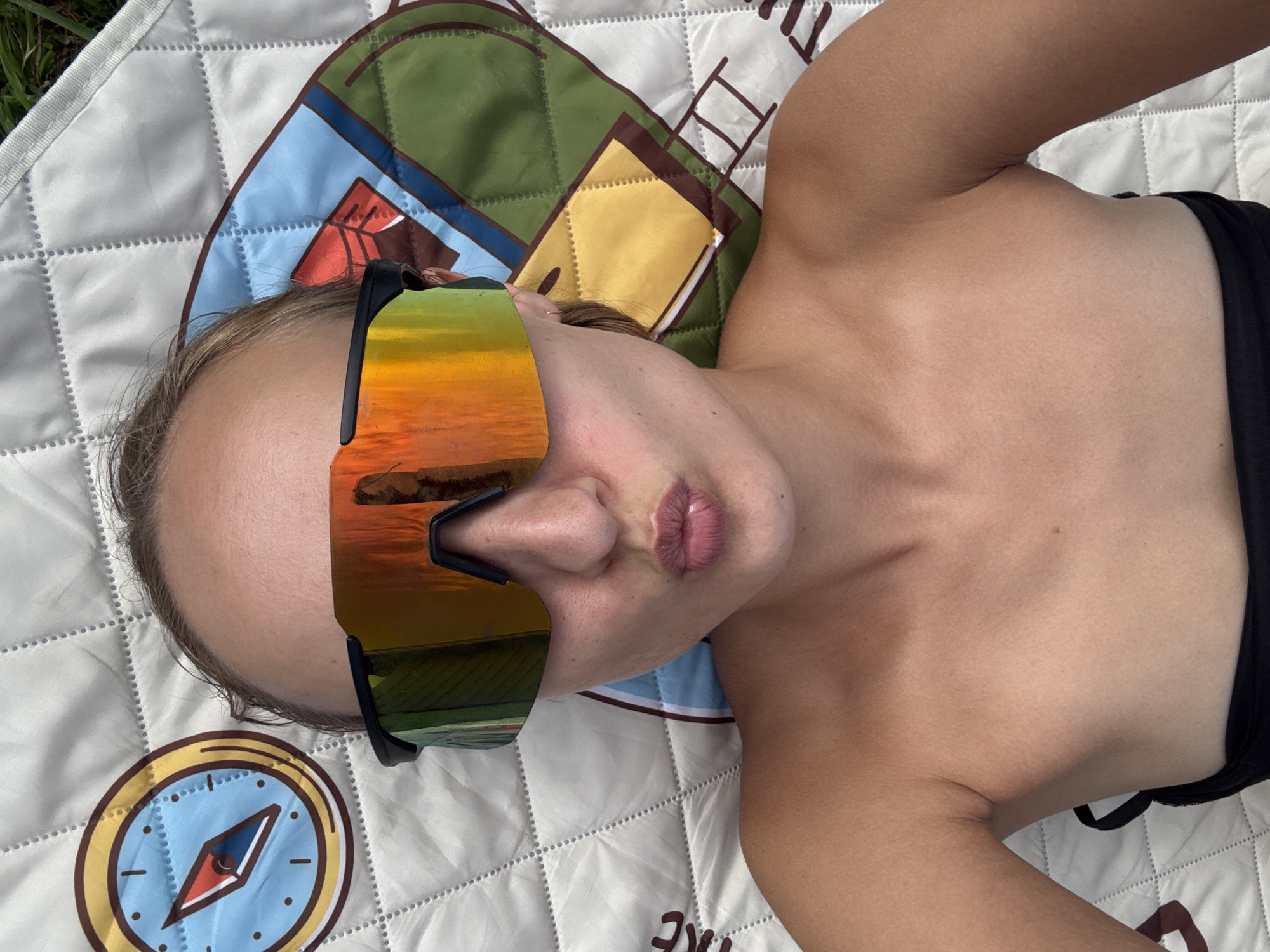 This is a top-down, vertical, close-up shot of a person lying on a white quilted picnic blanket patterned with an illustration of a compass. The person is wearing large, reflective sunglasses with orange and yellow lenses that show a reflection of a sunset over water, and black bottoms. The person's head is angled towards the left of the frame, with their face partially obscured by the sunglasses. Their skin appears tanned. The scene is lit by natural light, suggesting it is daytime, likely afternoon or early evening given the reflected sunset. The picnic blanket has a quilted diamond pattern and a border with a gray edge. The background is out of focus, showing green grass. The overall mood appears relaxed and summery. No visible text is present on the person or their attire, but the compass graphic on the blanket is a prominent design element. The location context provided is Подшивалово, Russia.