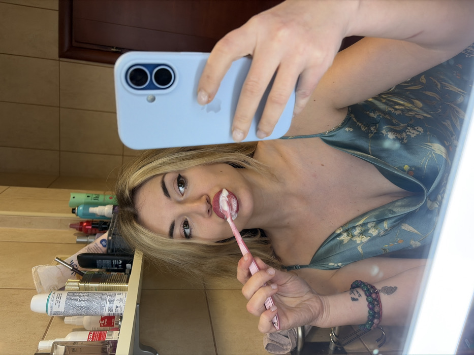The image displays a female individual taking a mirror selfie inside a bathroom. The location is specified as Budva, Montenegro. The woman has long, blonde hair and is depicted in the act of brushing her teeth with a pink toothbrush, which is covered in white foam around her mouth. Her gaze is directed towards her left (viewer's right). She is wearing a teal-colored satin-like slip dress with a floral pattern and thin shoulder straps. In her right hand, she holds a light blue iPhone, featuring two visible camera lenses and an Apple logo, positioning it to capture her reflection. Her left wrist displays multiple beaded bracelets, one with a small charm, and a tattoo is partially visible on her forearm. The bathroom background, visible in the mirror's reflection, consists of light brown tiled walls. A shelf situated above the mirror contains numerous personal care products, including bottles and aerosol cans. A dark brown wooden door is reflected in the upper right portion of the image, while a chrome towel rack with a light-colored towel is faintly visible on the left. Artificial light illuminates the scene.