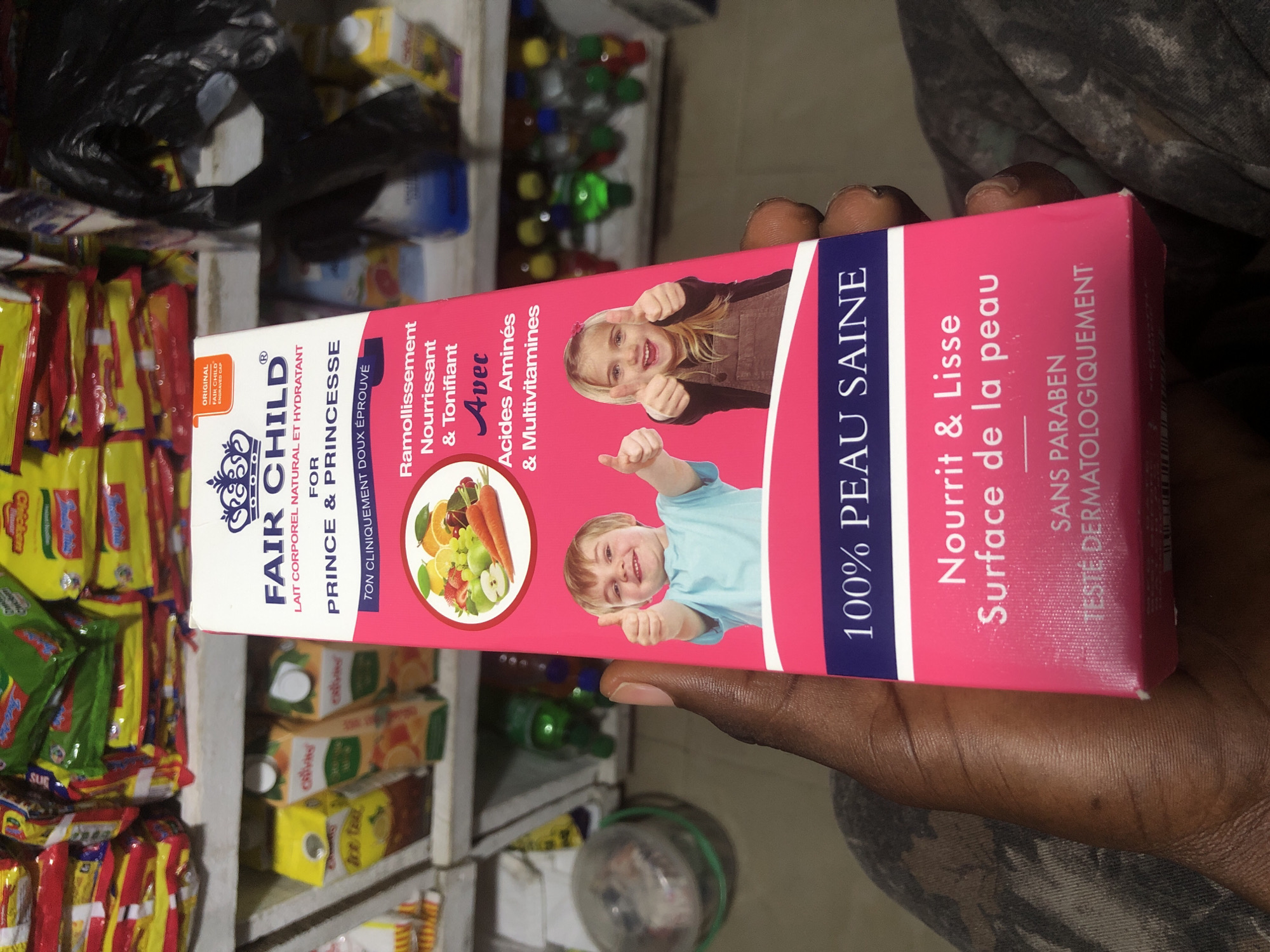A hand is holding a pink box of "FAIR CHILD" body lotion in a store. The box has images of children and fruit. The background shows shelves stocked with various products, including juice boxes and packaged snacks. The store appears to be in Ado, Nigeria.