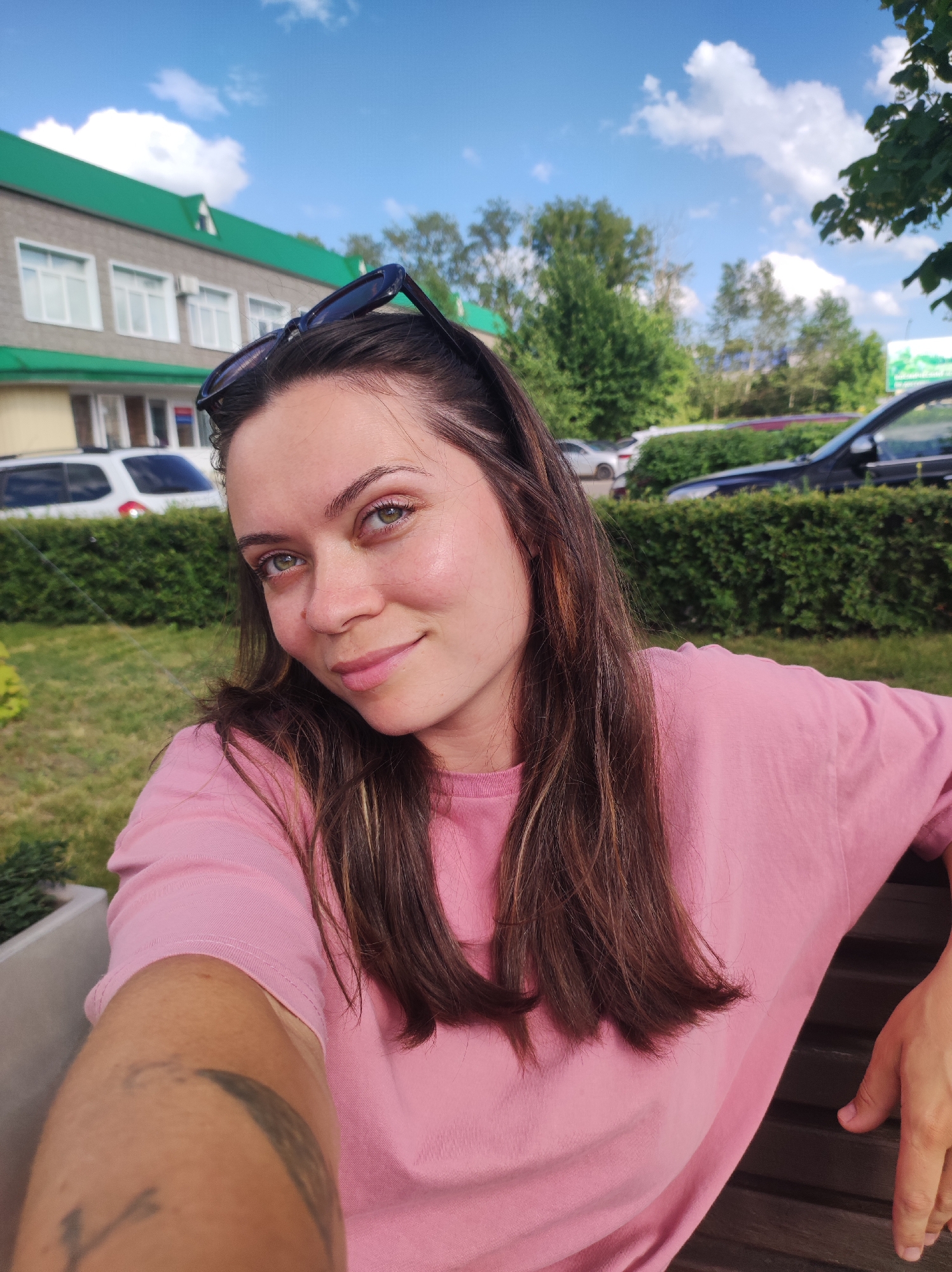 The media file is a selfie taken outdoors on a sunny day, likely in the afternoon given the bright lighting and clear blue sky with scattered clouds. The subject is a young woman with long, dark brown hair, wearing a pink t-shirt. She has a light tan and appears to be relaxed and happy, with a slight smile. She is sitting on a dark wooden bench, with her arm resting on it. Sunglasses are perched on top of her head.
In the background, there is a building with a green roof and grey walls, featuring several windows. Adjacent to the building is a line of green bushes, and behind that, cars are parked. The scene suggests an urban or suburban setting, possibly a public park or an area near a commercial building. The presence of trees further reinforces an outdoor environment. There is no visible text in the image. The overall mood is casual and pleasant.