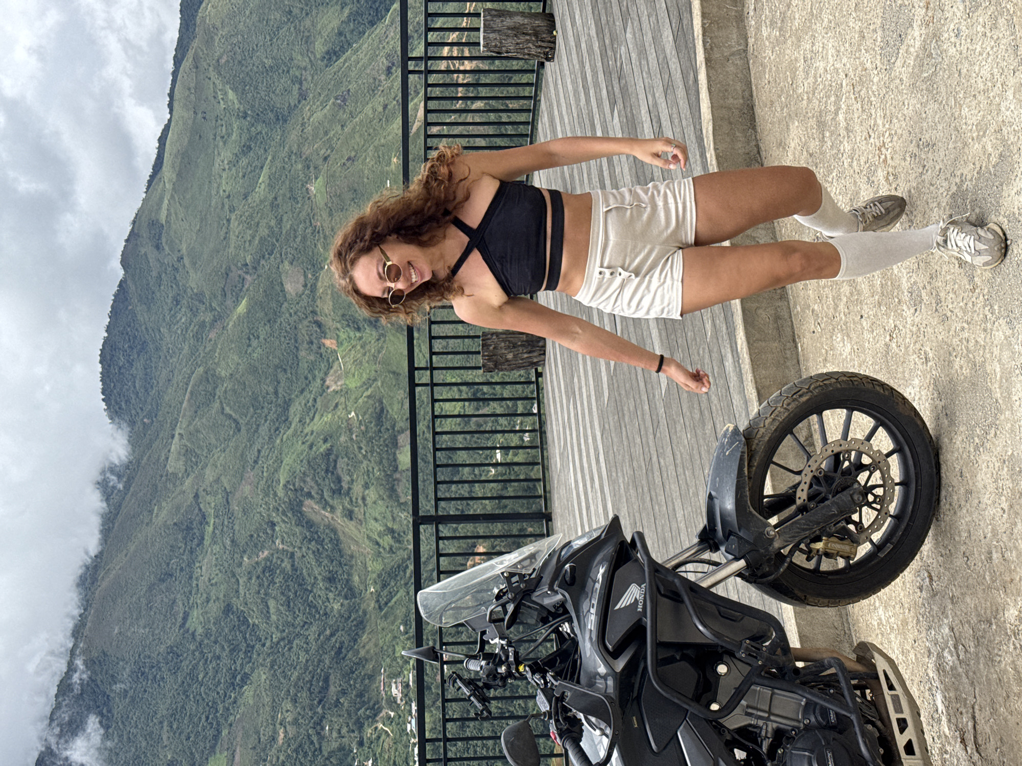 A young woman with curly brown hair, wearing sunglasses, a black halter-neck crop top, and light-colored shorts, stands on a wooden deck next to a black motorcycle. She is smiling and looking to her left. Behind her, a railing separates the deck from a steep, tree-covered mountainside under a cloudy sky. The location is identified as the city of Ta Xua, Vietnam.