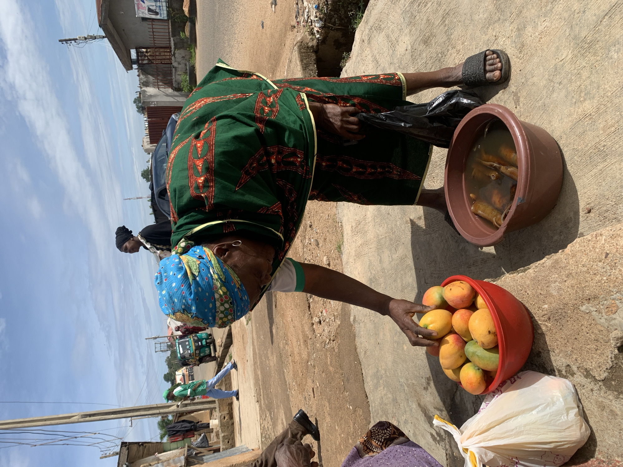 The image depicts an outdoor street vending scene in Jos, Nigeria, under a bright, cloudy sky. In the foreground, a dark-skinned woman, dressed in a multi-colored blue headwrap, a green patterned wrapper with yellow trim, and dark slides, bends over. Her right hand reaches into a red plastic bowl filled with ripe yellow, orange, and green mangoes placed on a concrete slab. Her left hand holds a black plastic bag. Adjacent to the mangoes, a brown plastic bowl contains several elongated, brown food items submerged in a clear liquid. A white plastic bag is visible to the left of the mango bowl.
Partially visible on the far left, another person's lower legs and feet, wearing dark shoes, are beside a purple garment, suggesting a customer or companion. In the middle ground, a man in a green shirt and blue trousers walks on a dirt path. Behind him, a green motorized tricycle taxi (keke napep) with "GIFT FROM GOD" printed on its front is parked. Further right, a woman in a black headwrap and dark attire stands observing the scene.
The environment includes a dusty dirt road running parallel to the concrete pavement. On the right, a single-story concrete building with a partial sign displaying "DAYCARE" and "NURSERY" is visible behind a low metal fence. Another dilapidated building stands on the far left. Two utility poles with multiple overhead wires are present: a tall, slender pole on the left and a shorter pole with a junction box on the right. Scattered debris is visible on the ground near the fence on the right. The sky overhead is light blue with dispersed white clouds.