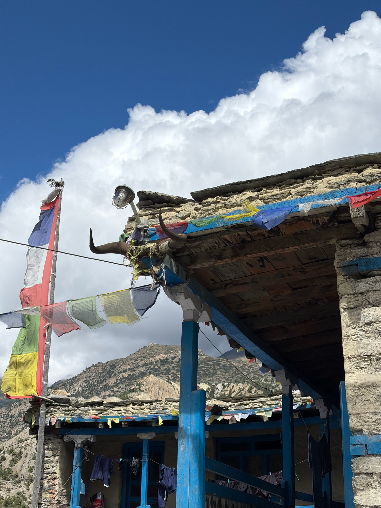 In the village of Ngawal, Nepal, a traditional stone and wood building stands against a backdrop of mountains and a partly cloudy blue sky. The structure features a weathered, multi-layered roof. Prayer flags in various colors, including blue, yellow, red, green, and white, are strung across the building's facade and from a flagpole. A pair of bull horns are mounted above an entranceway, adorned with more prayer flags and some yellow tassels. A metal lamp with a glass cover is affixed to a post near the horns. Laundry is hung out to dry on lines strung between blue-painted pillars in the foreground. The mountainous terrain behind the building is dry and rocky, with sparse vegetation.