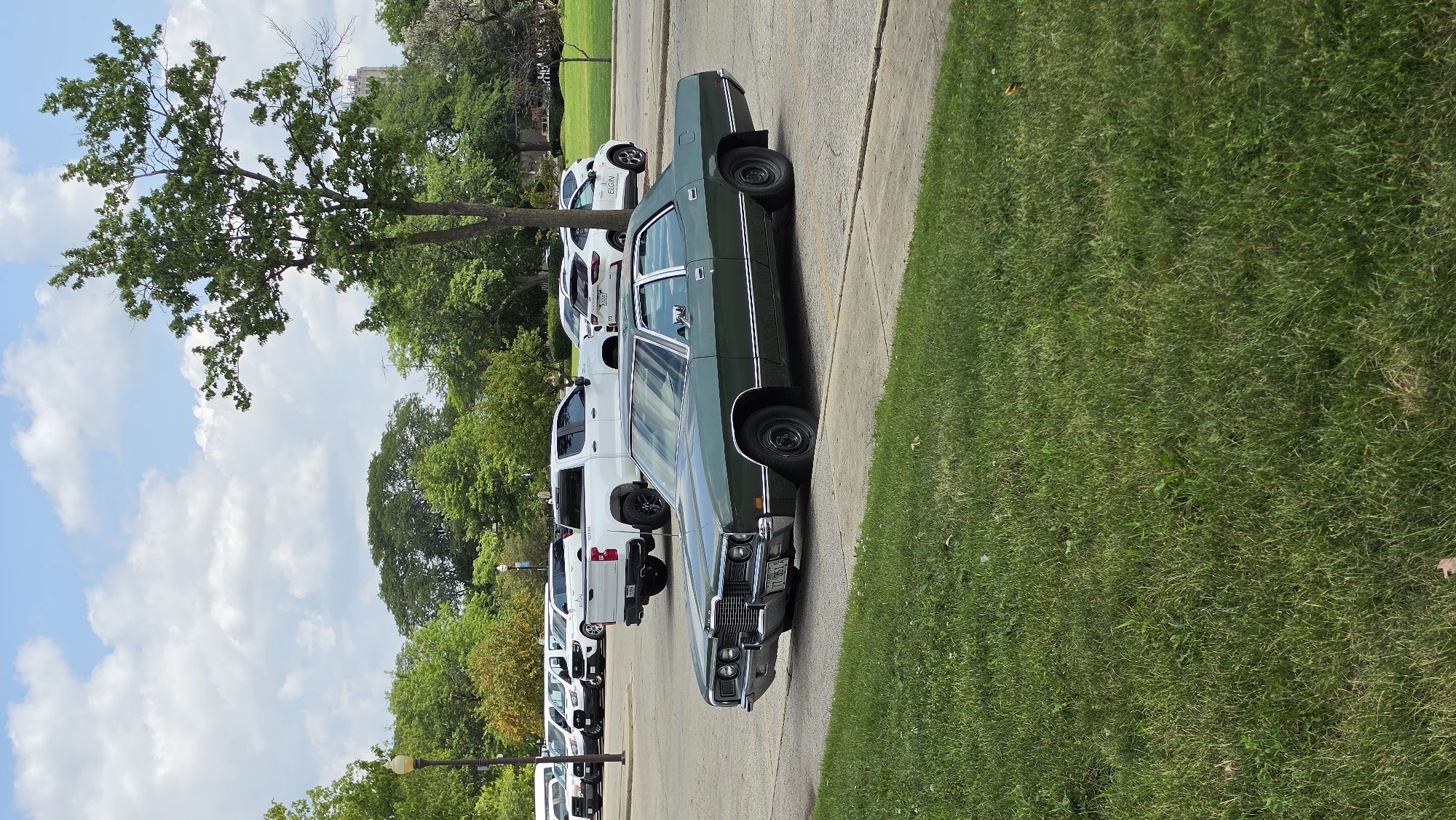 A green 1970s sedan is parked in a parking lot in Elgin, United States of America, with a white pickup truck and other vehicles in the background. The car is a Ford Galaxie 500. The front of the car has a chrome bumper and grille. The vehicle is facing the viewer, slightly angled to the right. The car is parked on asphalt, and there is a patch of green grass in the foreground. The sky is blue with scattered clouds. The scene appears to be a sunny day.
