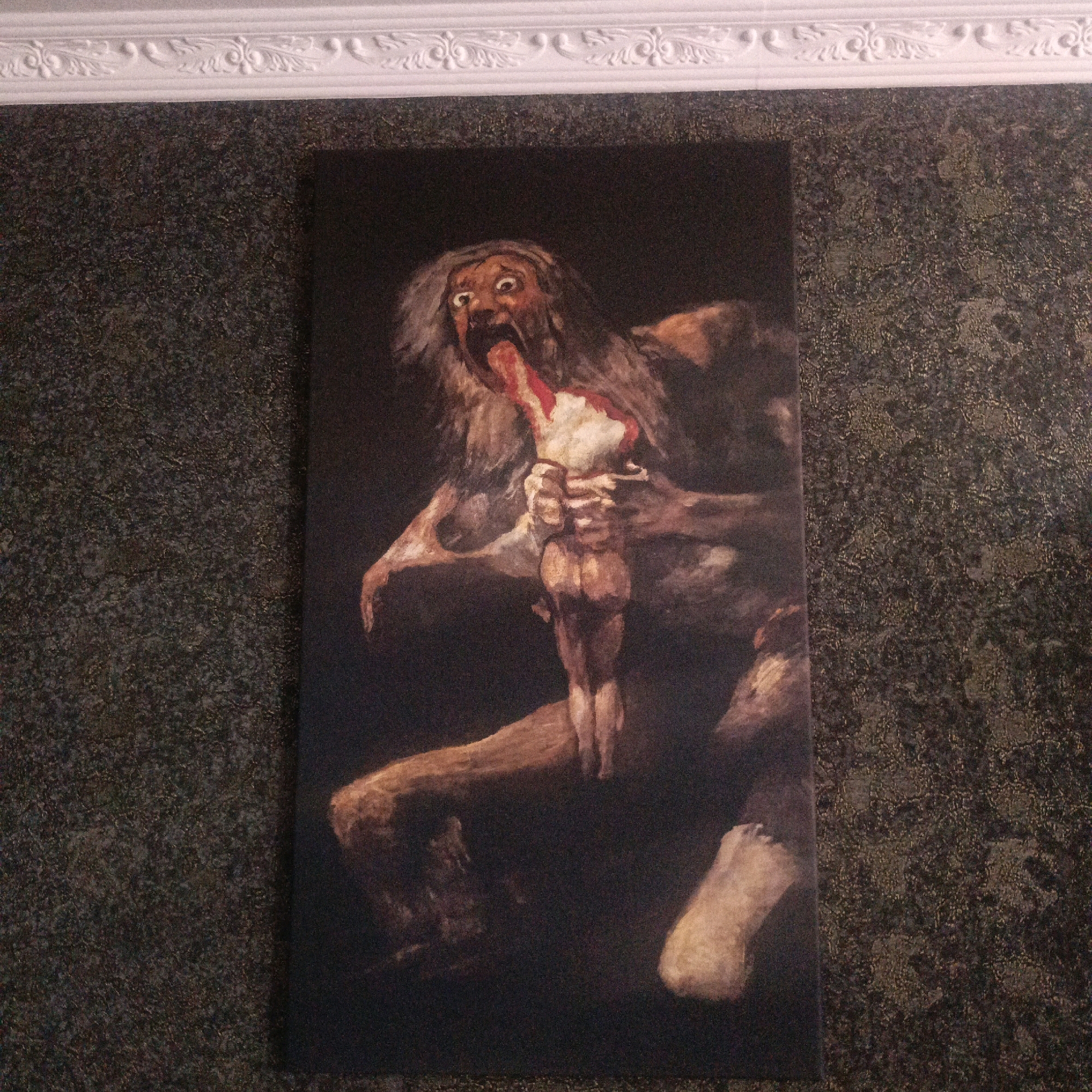 The image shows a painting of a monstrous figure, likely a depiction of Saturn devouring his son, in a dark, textured room. The painting is on a dark background, and the figure is rendered in a grotesque and violent manner, with sharp teeth, wild eyes, and a bloody tongue. The figure is tearing into a human-like form, with limbs and flesh visible. The overall impression is one of horror and despair. The painting is hung on a wall with a dark, textured wallpaper and a white decorative molding at the top. The exact location in Pervomayskiy, Russia is not specified, but it appears to be an indoor setting, possibly a home or a gallery.